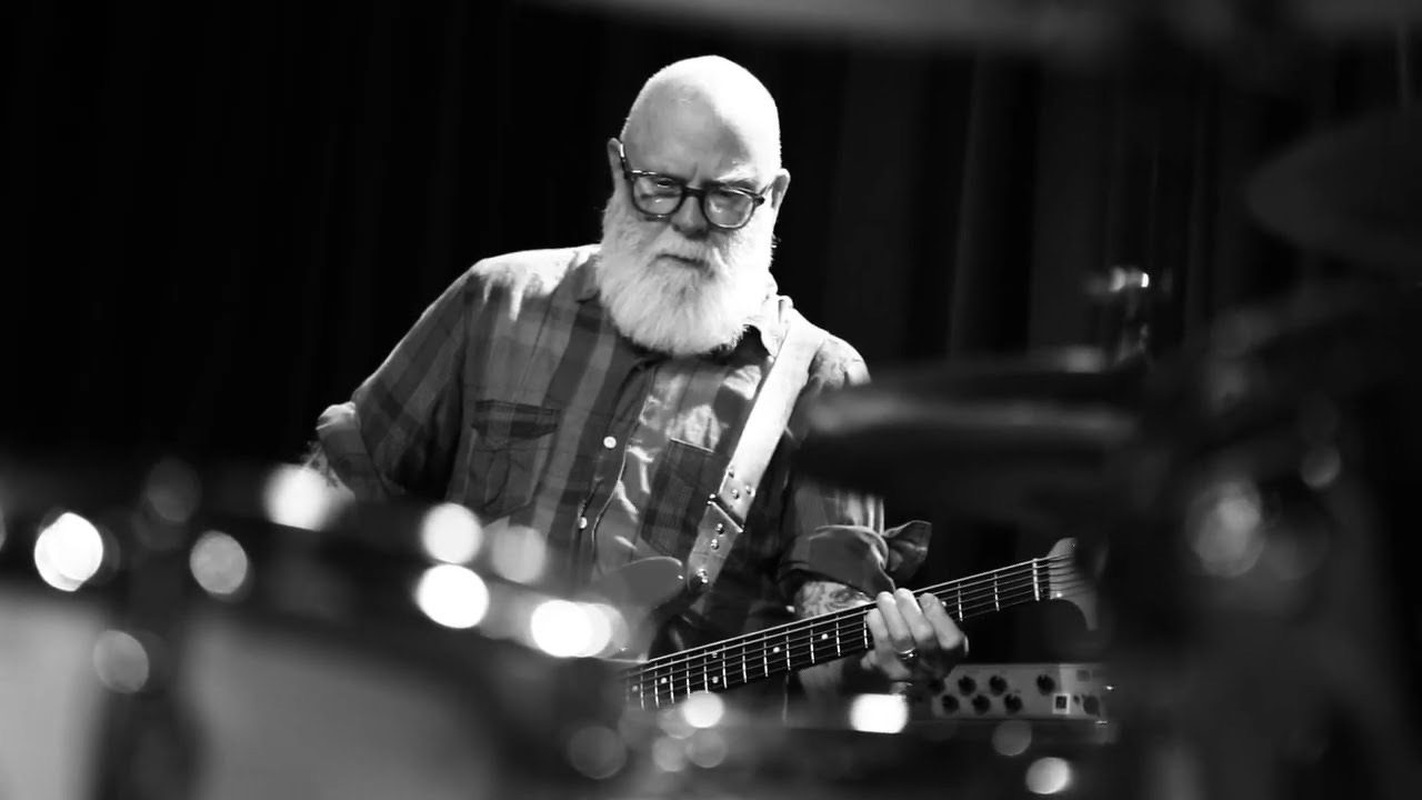 An older man with a white beard and glasses is playing an electric guitar on stage.