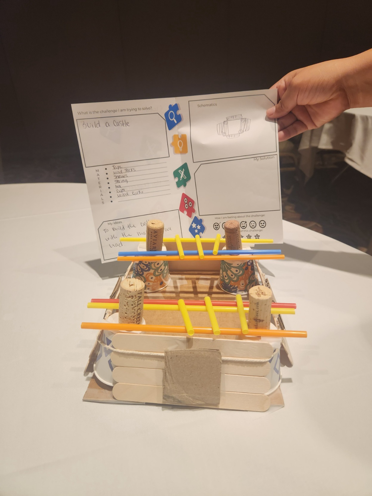 Auto-generated description: A handmade craft project resembling a small castle made from popsicle sticks, corks, and straws is accompanied by a sketched design plan on paper held by a hand.