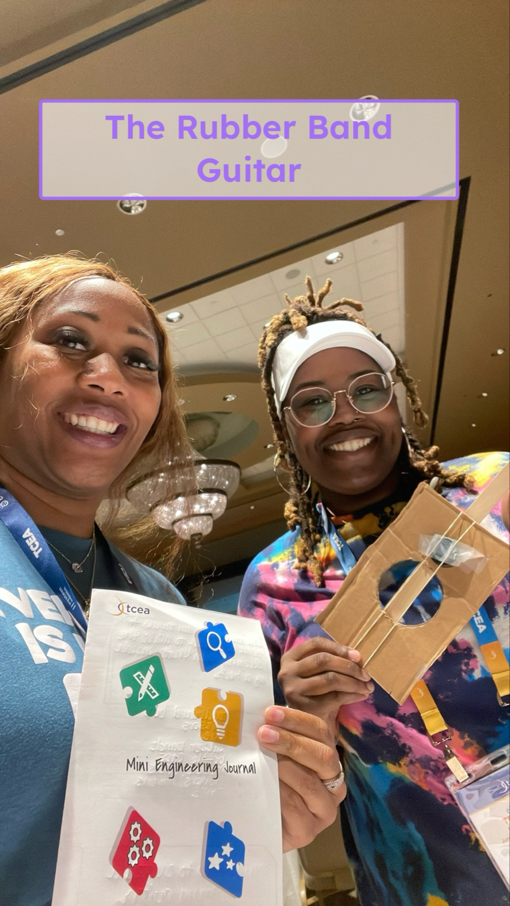 Auto-generated description: Two smiling individuals are holding up a Mini Engineering Journal and a cardboard rubber band guitar.