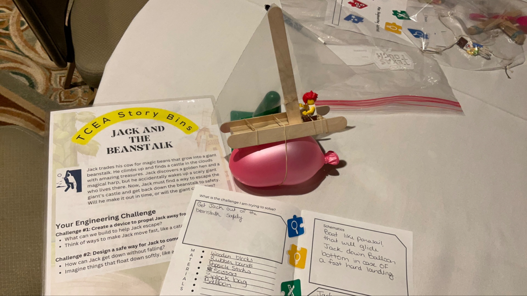 Auto-generated description: A table displays a STEM activity setup involving a Jack and the Beanstalk themed engineering challenge, with a plastic bag, instruction papers, and a balloon supporting a makeshift wooden structure with a toy figure on top.