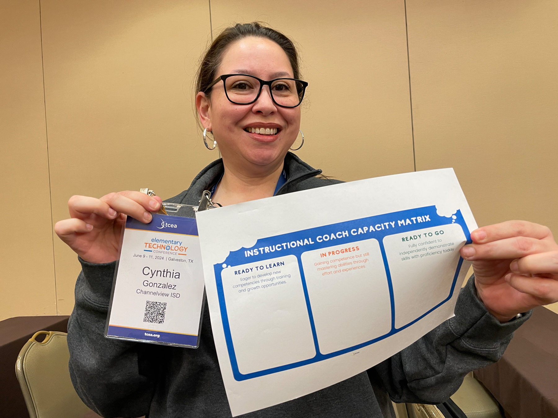 Auto-generated description: A person wearing glasses and holding a paper titled Instructional Coach Capacity Matrix is smiling and showcasing an ID badge labeled Cynthia Gonzalez.