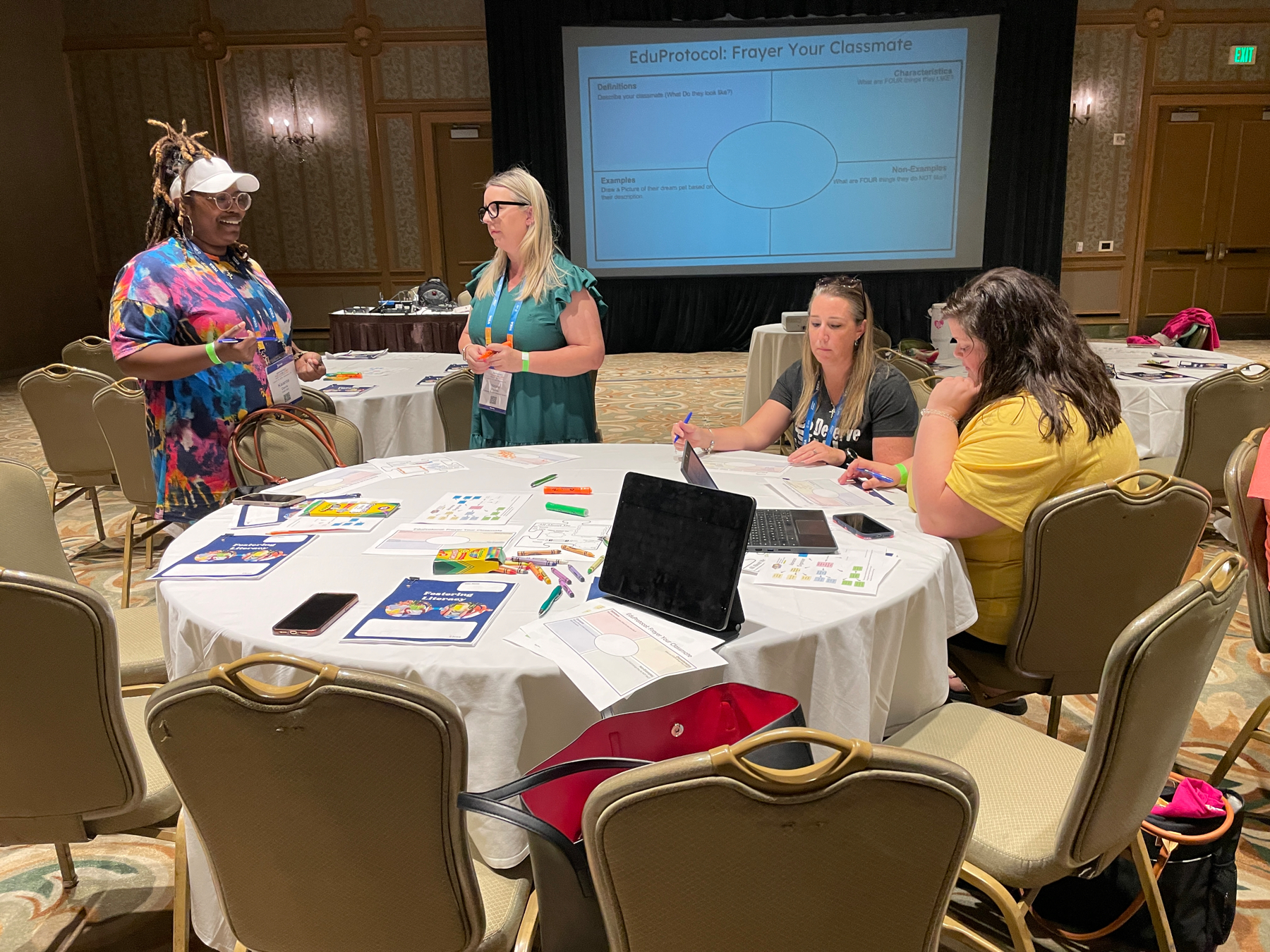 Auto-generated description: A group of people are engaged in discussion and activities around a table in a conference or workshop setting, with educational materials and a presentation screen in the background.