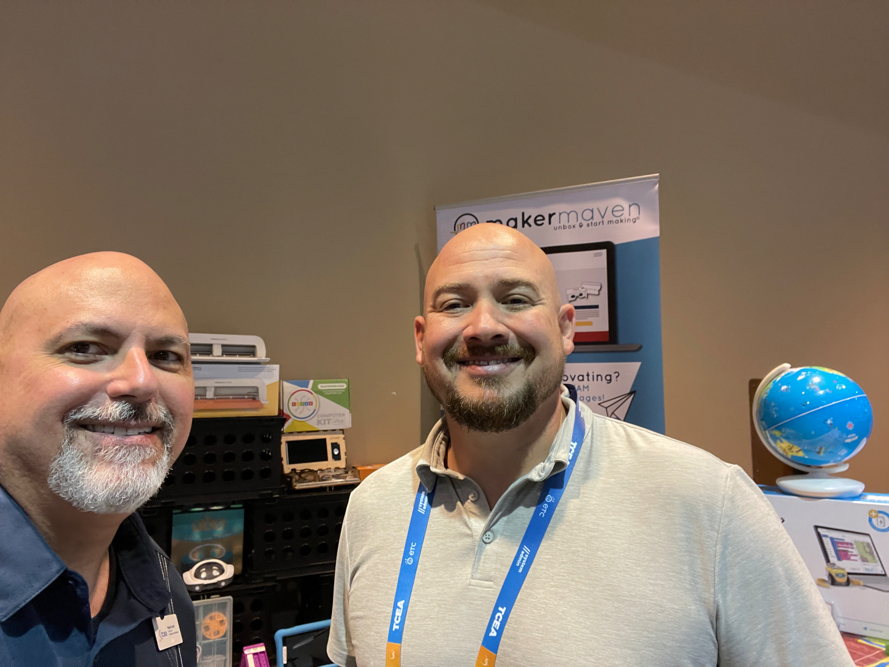 Auto-generated description: Two men (Miguel and Matt) are smiling with a display booth and a globe visible in the background.