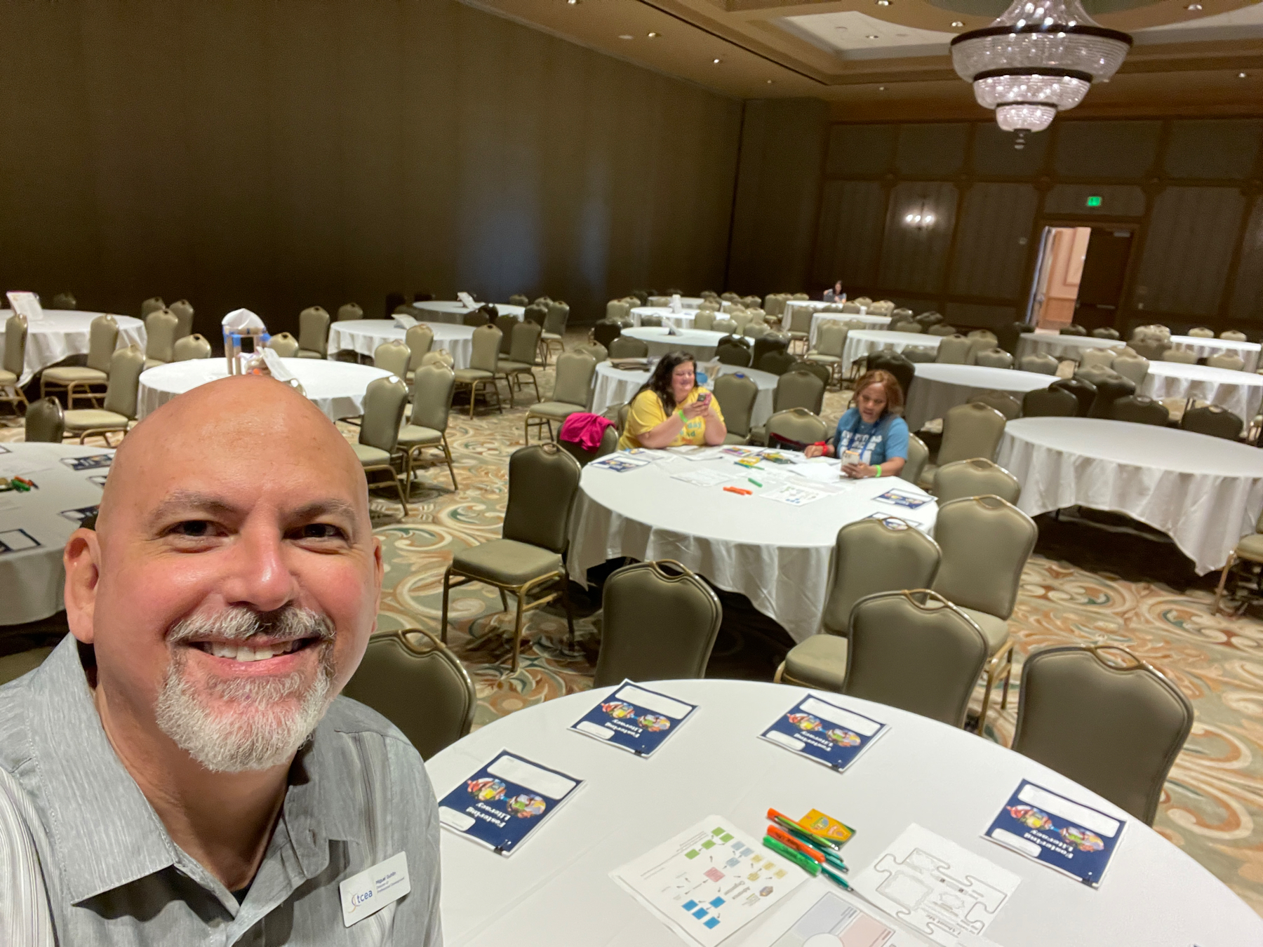 Auto-generated description: A man is taking a selfie in a conference room with empty round tables, while two people sit at one of the tables in the background.