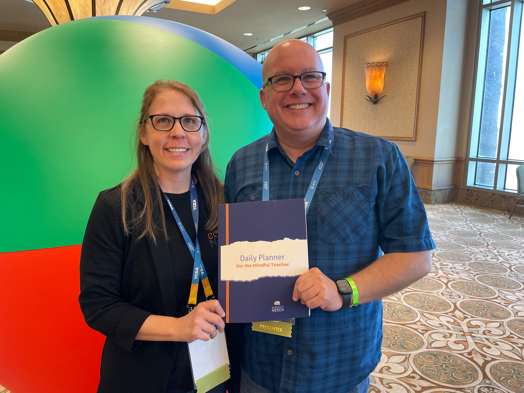 Auto-generated description: Two people, one holding a Daily Planner for the Special Teacher, are smiling and standing in front of a large colorful display ball in what appears to be a conference or event setting.