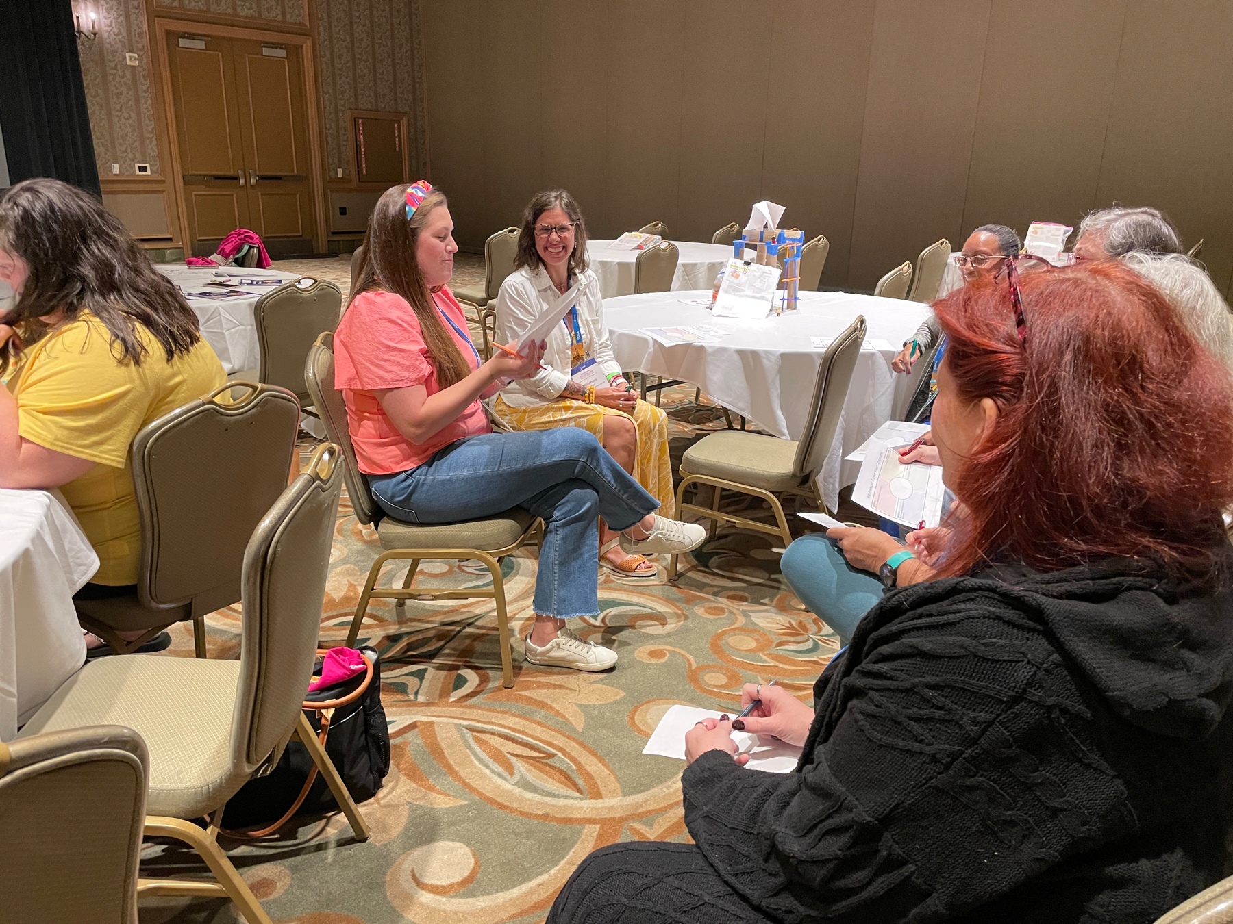 Auto-generated description: A group of people sits around a table in a conference room, engaging in conversation.