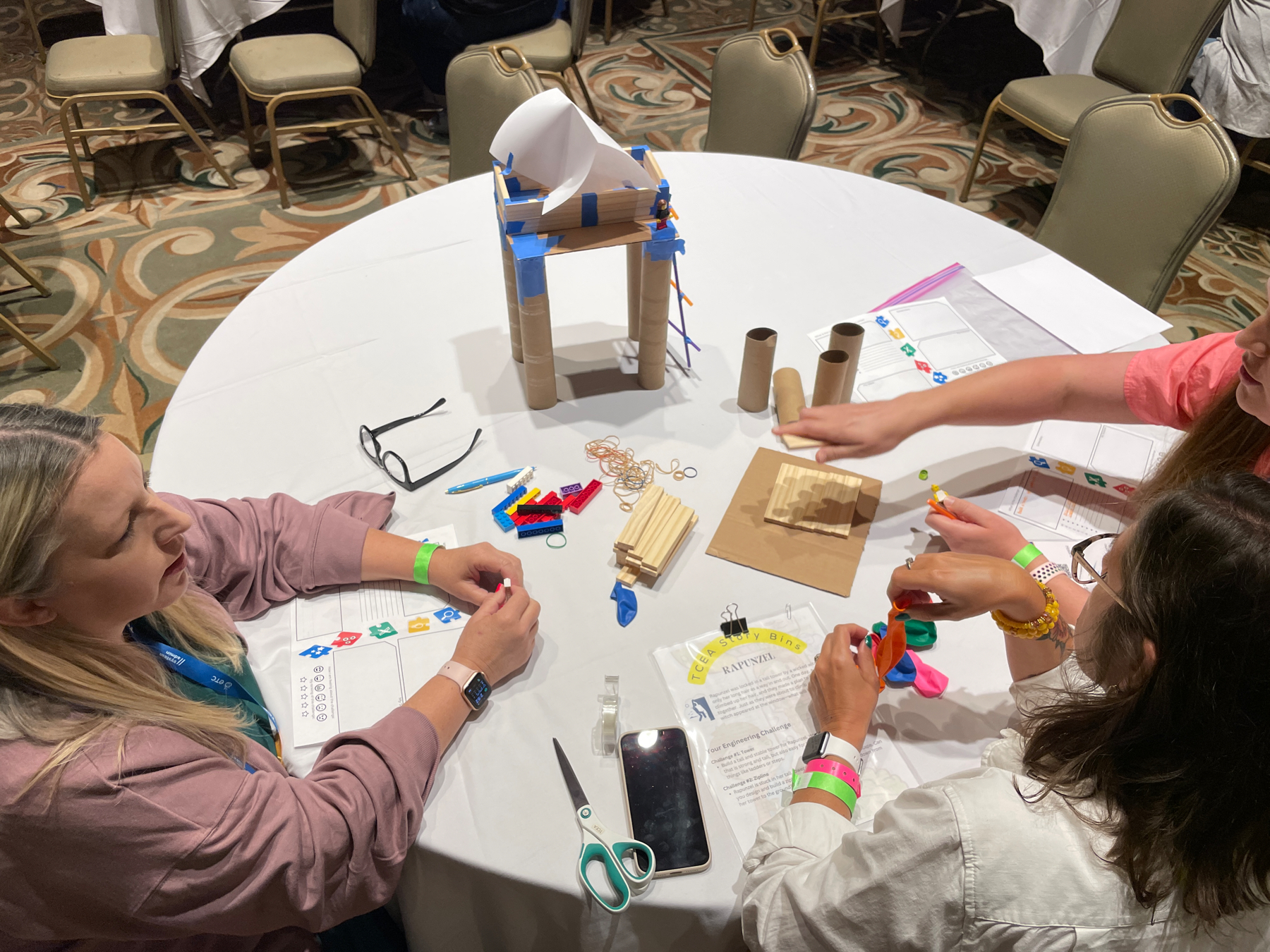 Auto-generated description: Three individuals engage in a hands-on activity at a round table, working on a project involving various crafting materials such as cardboard, tape, and scissors.