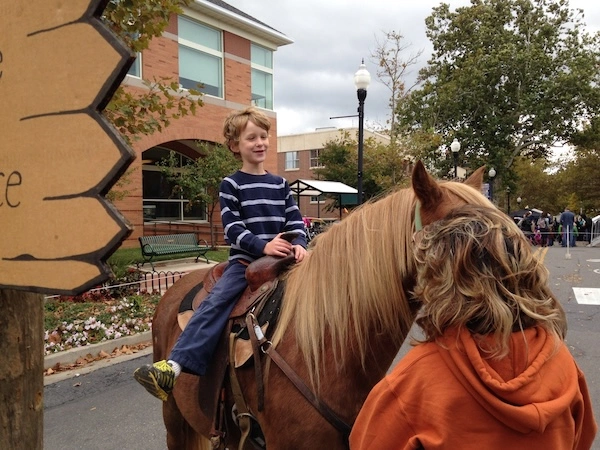 A boy wearing a striped shirt is happily riding a horse on a tree-lined street with brick buildings and buildings, accompanied by someone in an orange hoodie.