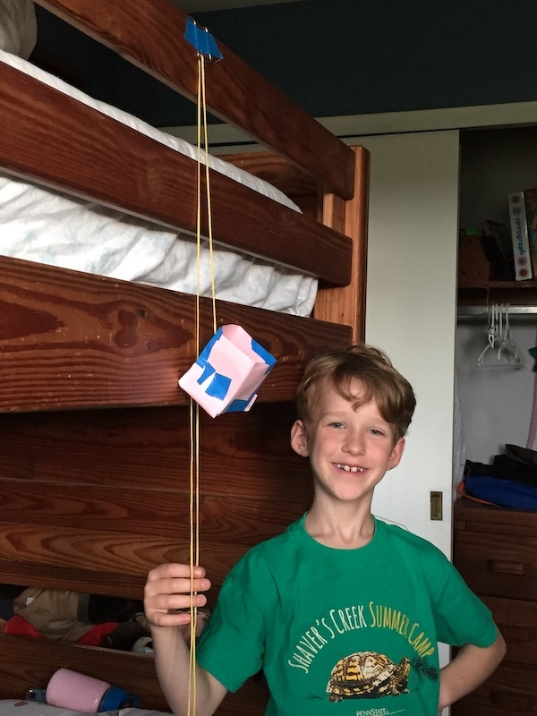 Auto-generated description: A smiling child is standing near a bunk bed, holding a string attached to a small, cube-shaped object covered in paper and tape.