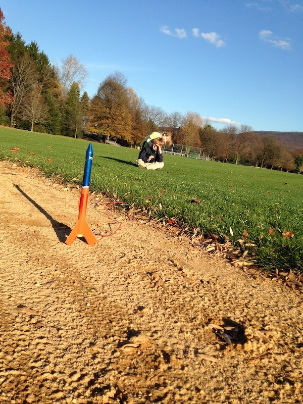 Auto-generated description: A small model rocket is ready for launch on a sandy patch, with grassy fields and trees in the background.