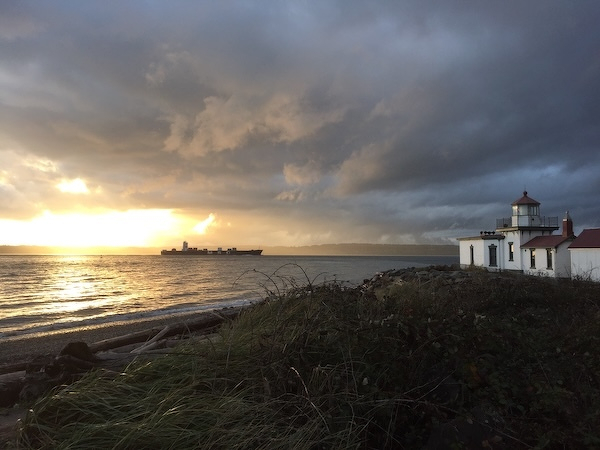 Auto-generated description: A lighthouse stands by a rocky shoreline under dramatic storm clouds, with a ship visible on the horizon as the sun begins to set.