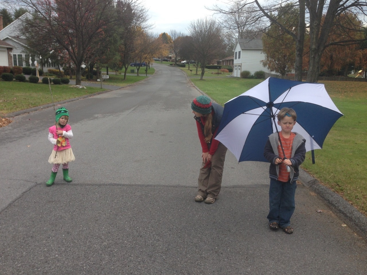 Two children, one holding a large umbrella, the other holding a stick, pause with their mother walking a suburban street