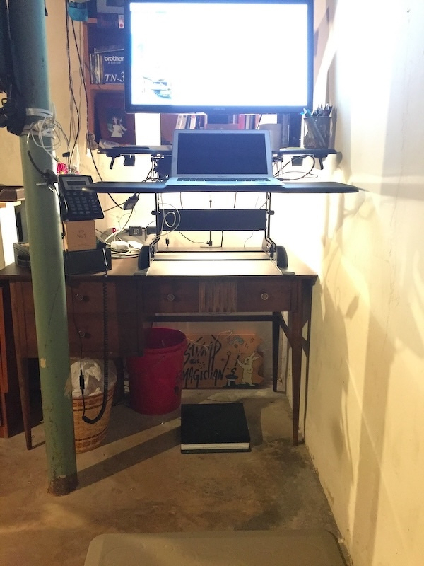  A makeshift standing desk setup is placed on a wooden table in a basement, featuring a laptop, a monitor, and various desk accessories.