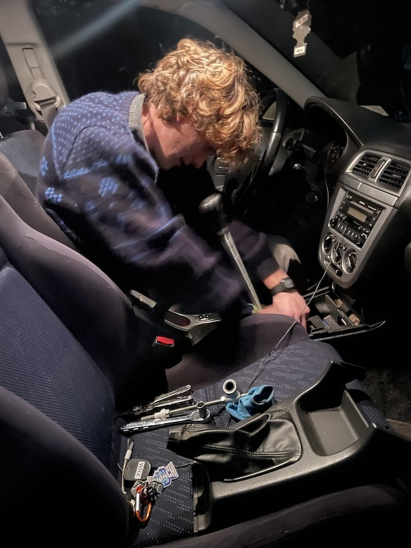 A person wearing a patterned sweater works on the center console of a car interior, with tools and car keys on the seat.