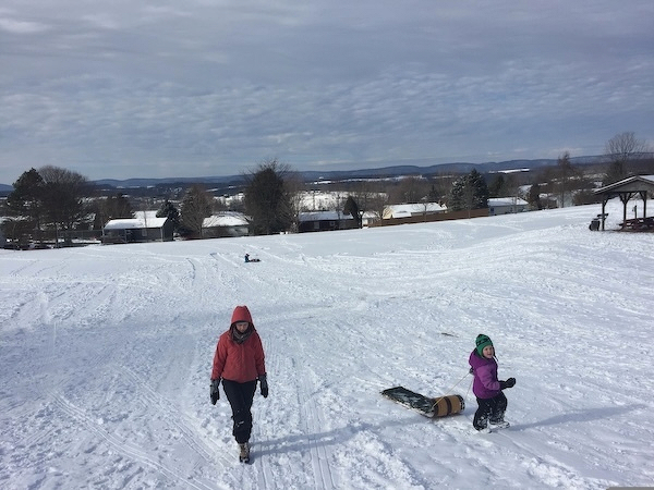 Three people in on a snowy landscape: a woman walking up a hill, a little girl walking up the same hill pulling a toboggan, and someone at the bottom of the sledding hill with a snow tube.