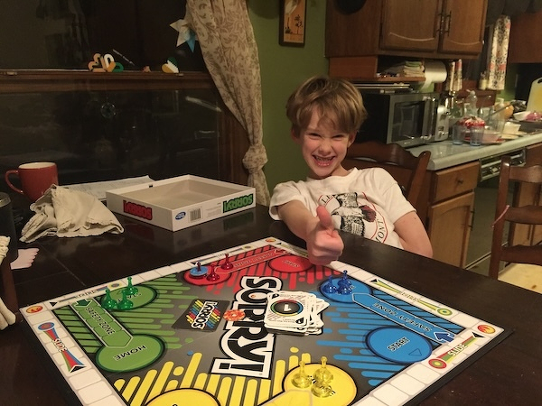 A smiling boy gives a thumbs-up while sitting at a table with a colorful board game.
