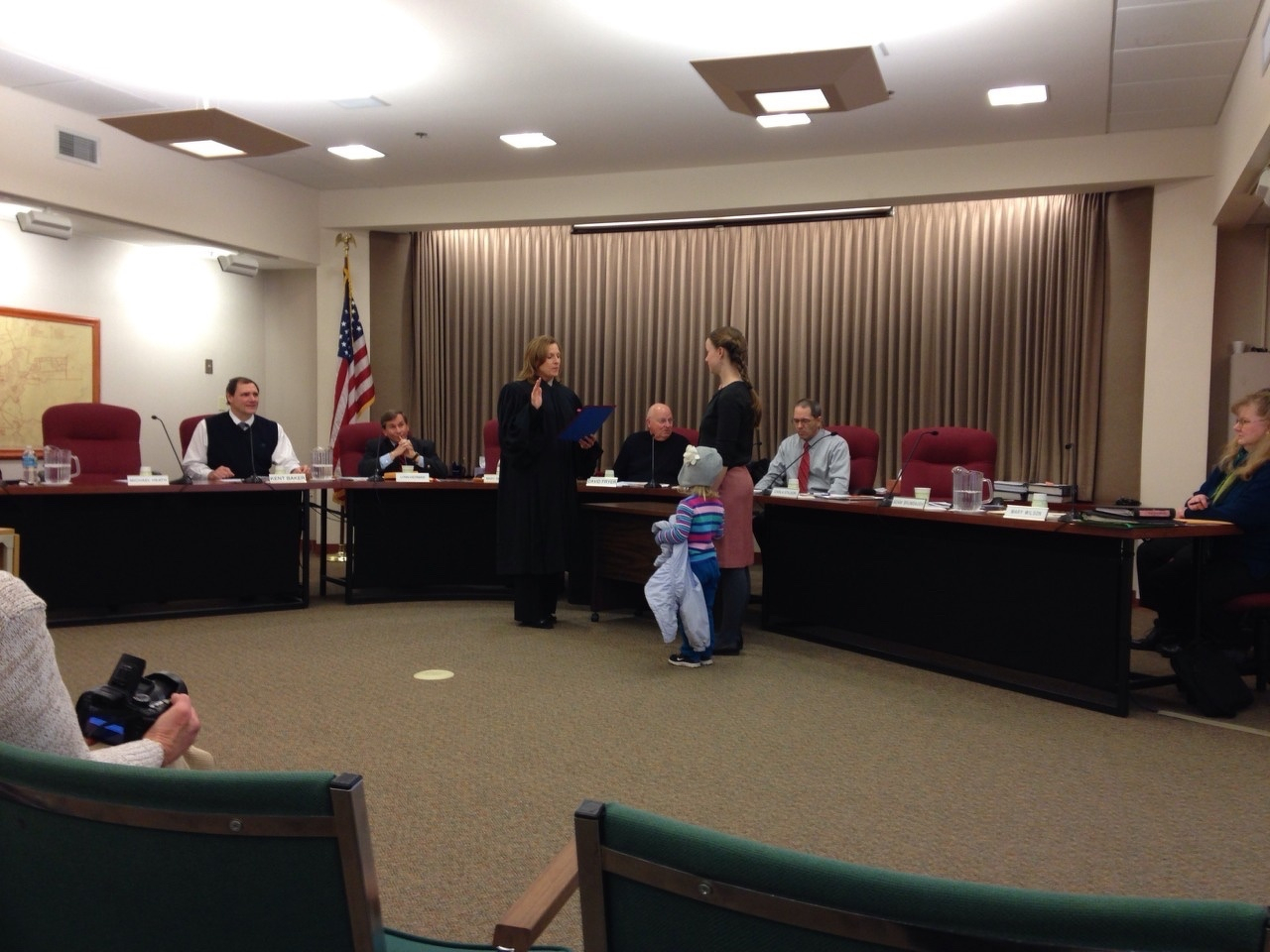  A formal meeting or ceremony is taking place in a courtroom-style setting, where a judge or official is addressing a woman with a child at her side, with other officials seated at a long table.