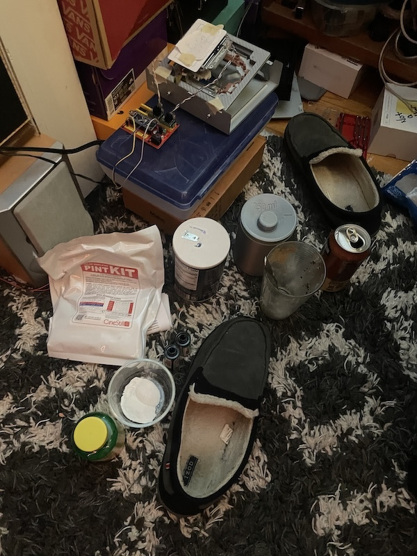 A cluttered floor features various household items, including slippers, containers, a can, and electronic components on a rug.