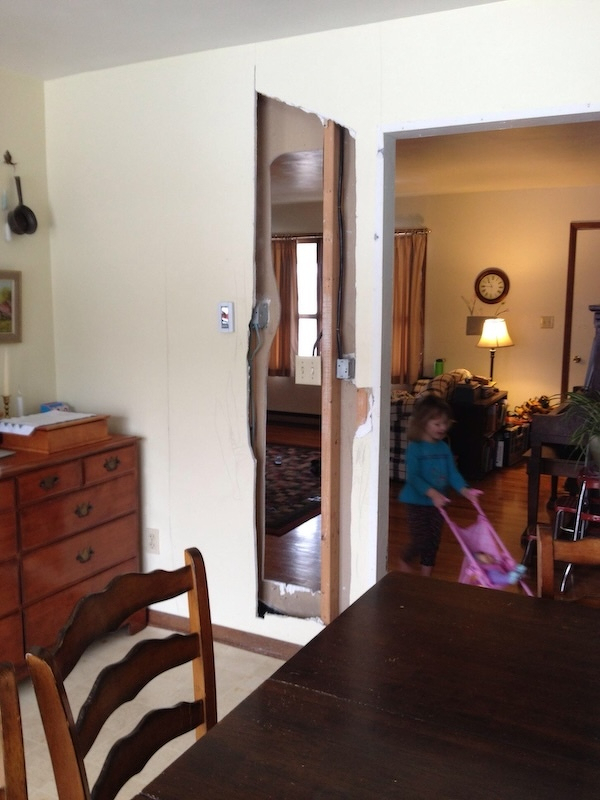 A child pushes a toy stroller through a room with a partially removed wall section, revealing exposed wooden studs.