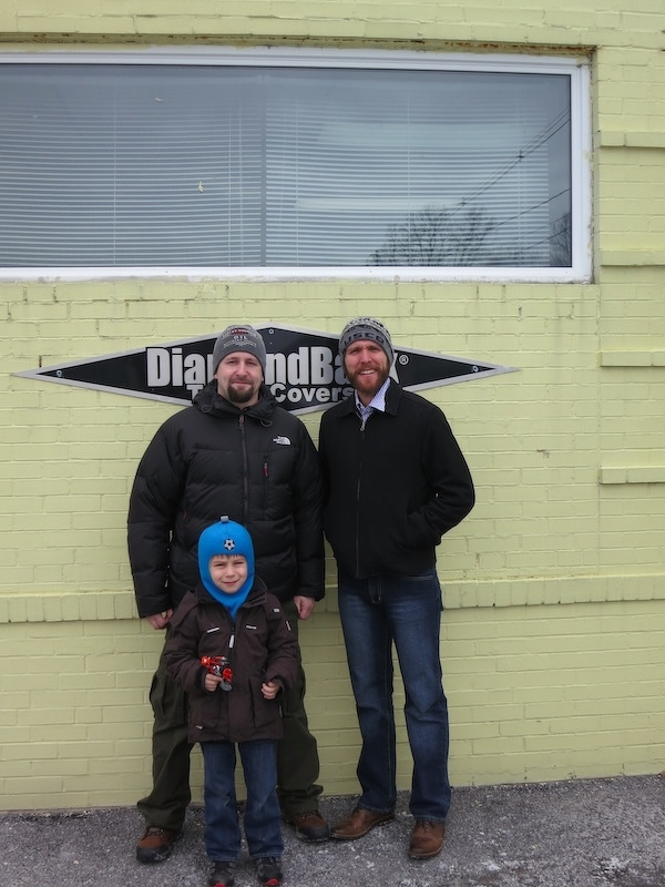 Three people, including a child holding a toy, are standing outside in front of a yellow brick building.