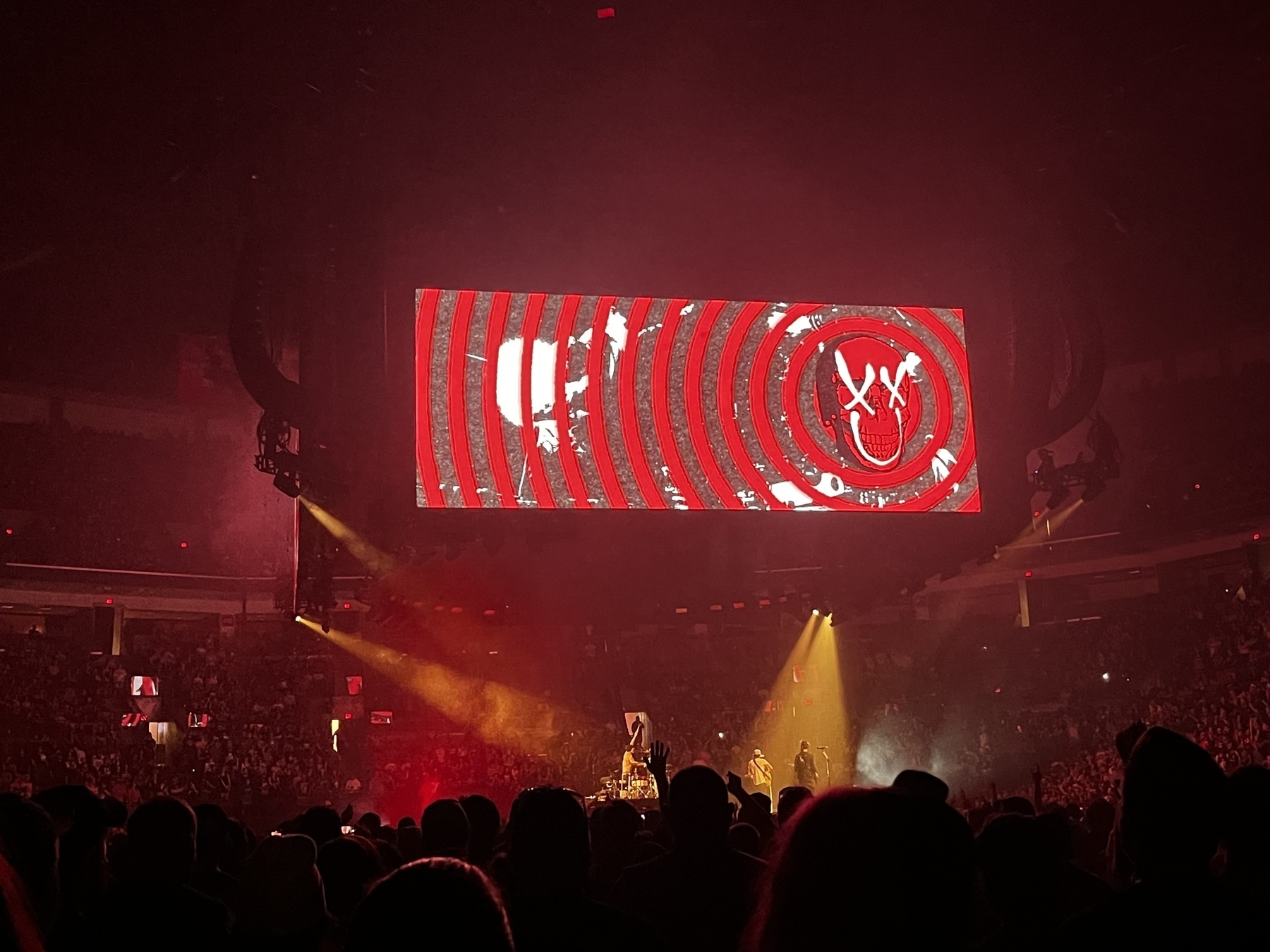 Auto-generated description: A crowd watches a concert with a stage illuminated by red and yellow lights and a large screen displaying circular patterns and symbols.