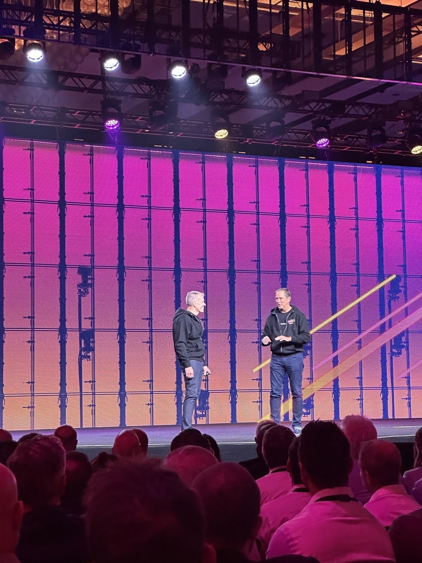 Two individuals are standing on a stage with a large screen behind them, speaking to an audience.