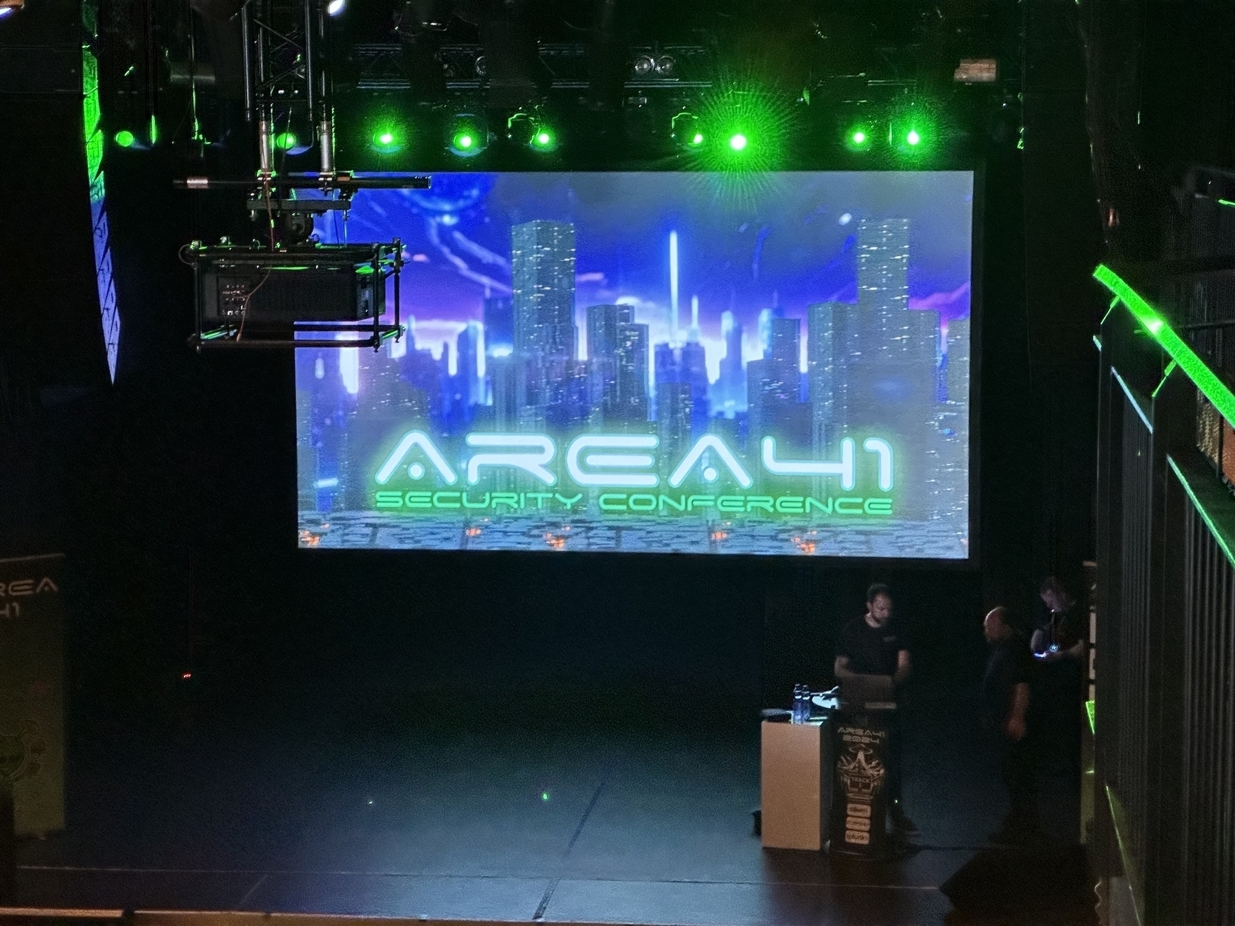 A conference stage is set up with a large screen displaying AREA41 SECURITY CONFERENCE and futuristic cityscape graphics, accompanied by green lighting and people preparing on the side.