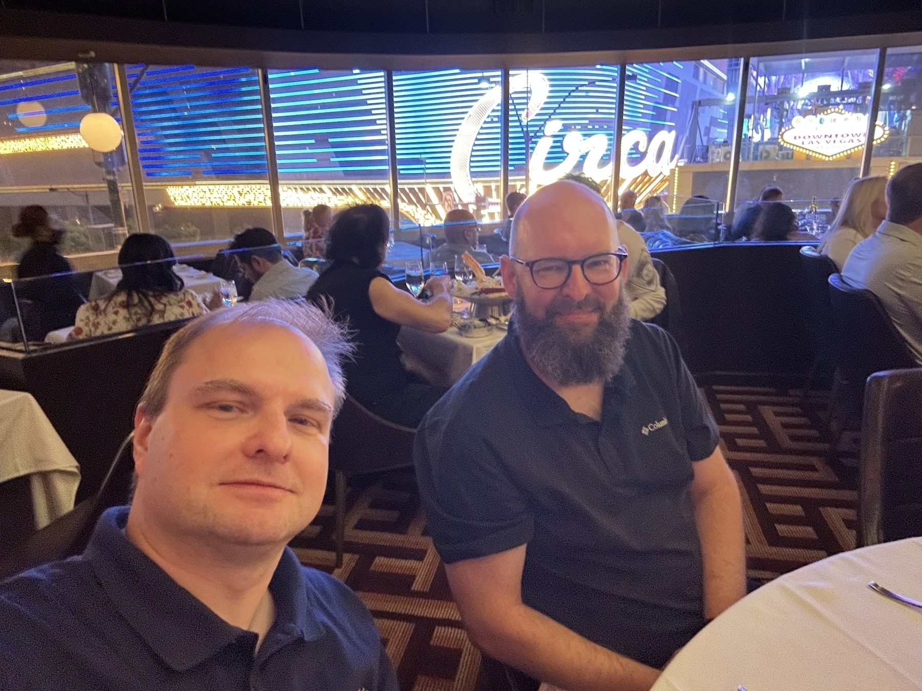 Two men are sitting at a table inside a bustling restaurant with a large illuminated Circa sign visible through the window behind them.