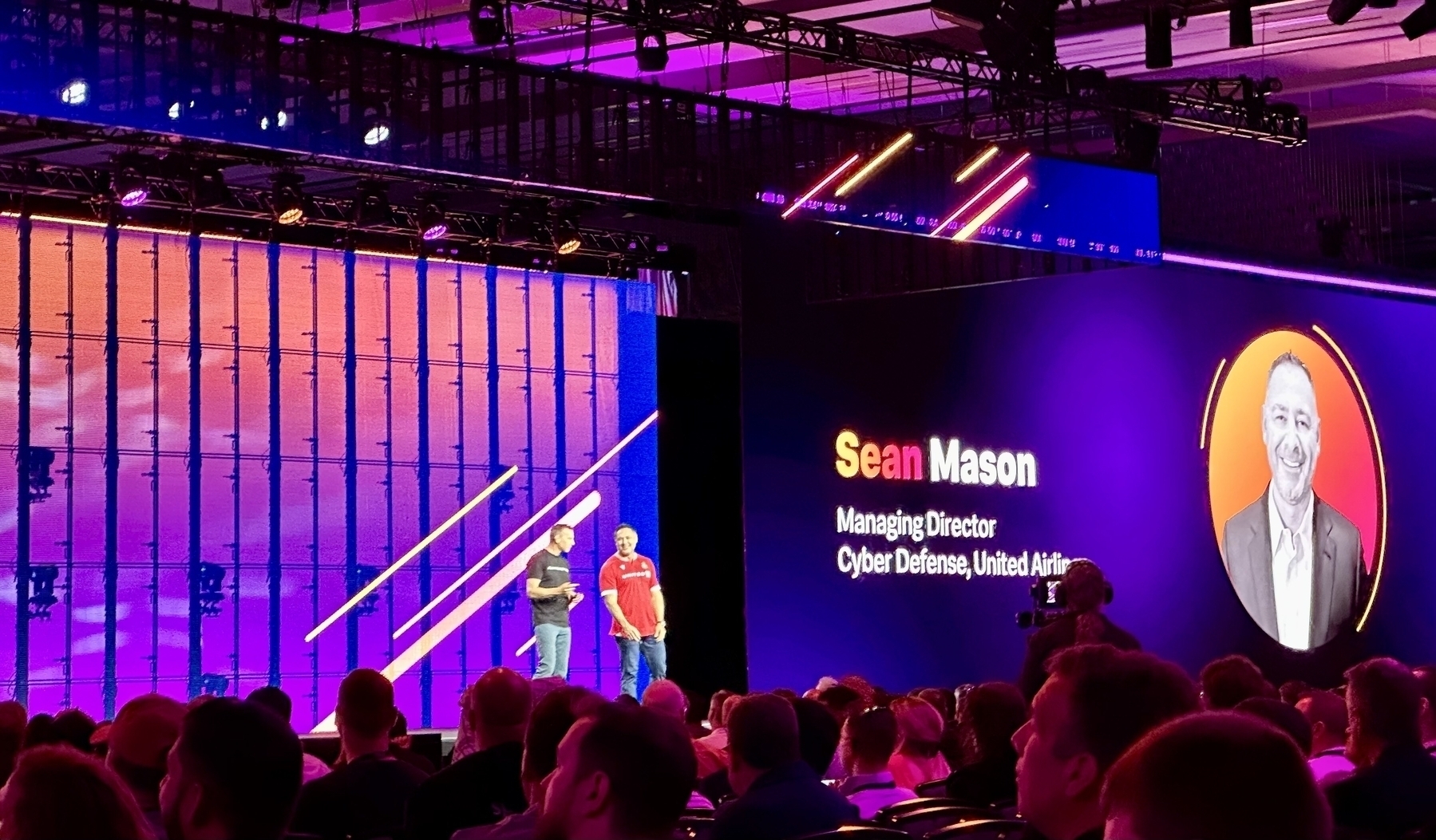 Two people are standing on a stage in front of an audience, with a large screen displaying the name Sean Mason and his professional title.