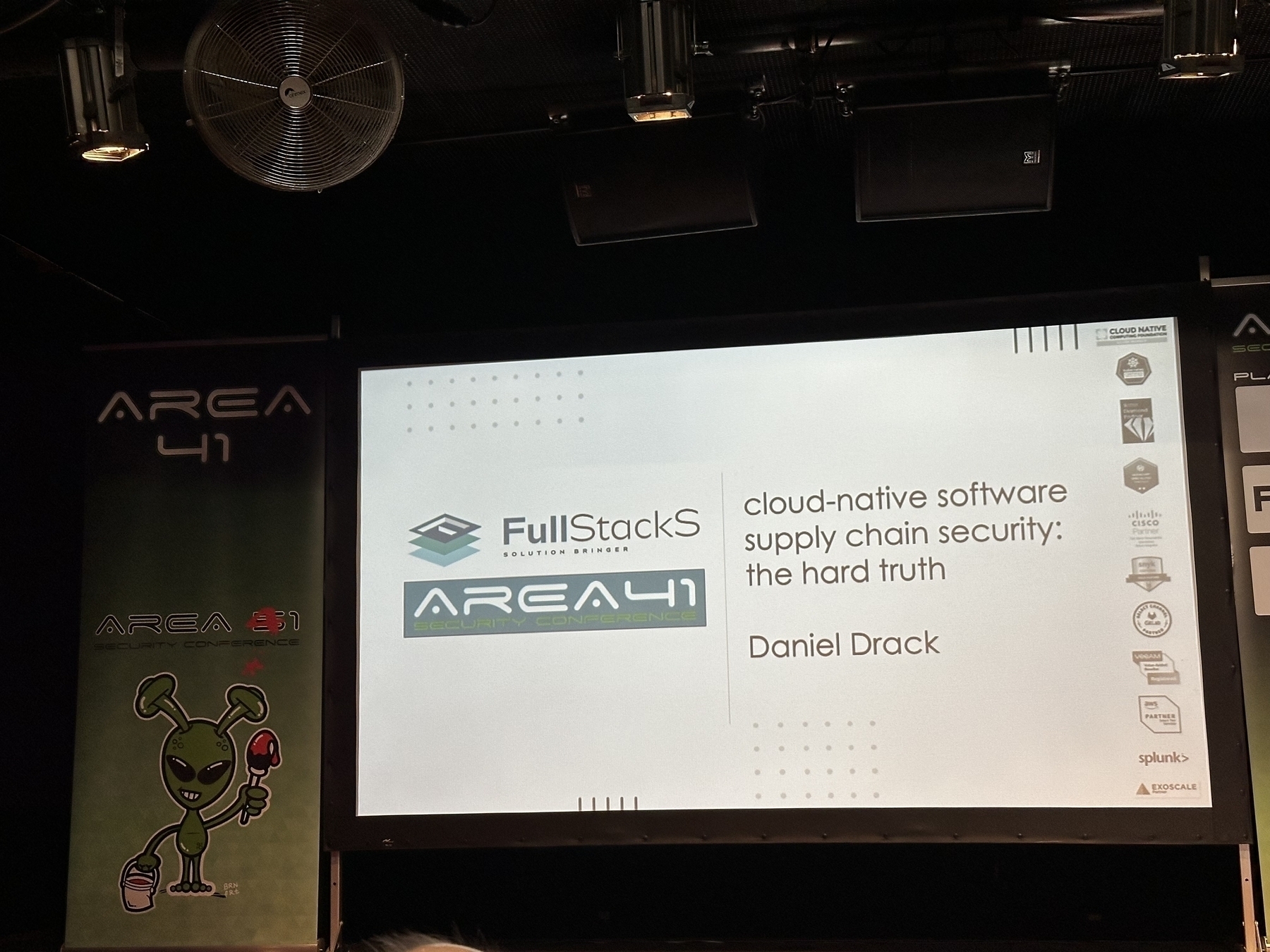 A conference presentation slide on a screen shows details about cloud-native software supply chain security by Daniel Drack, with an Area 41 banner and an alien illustration in the background.