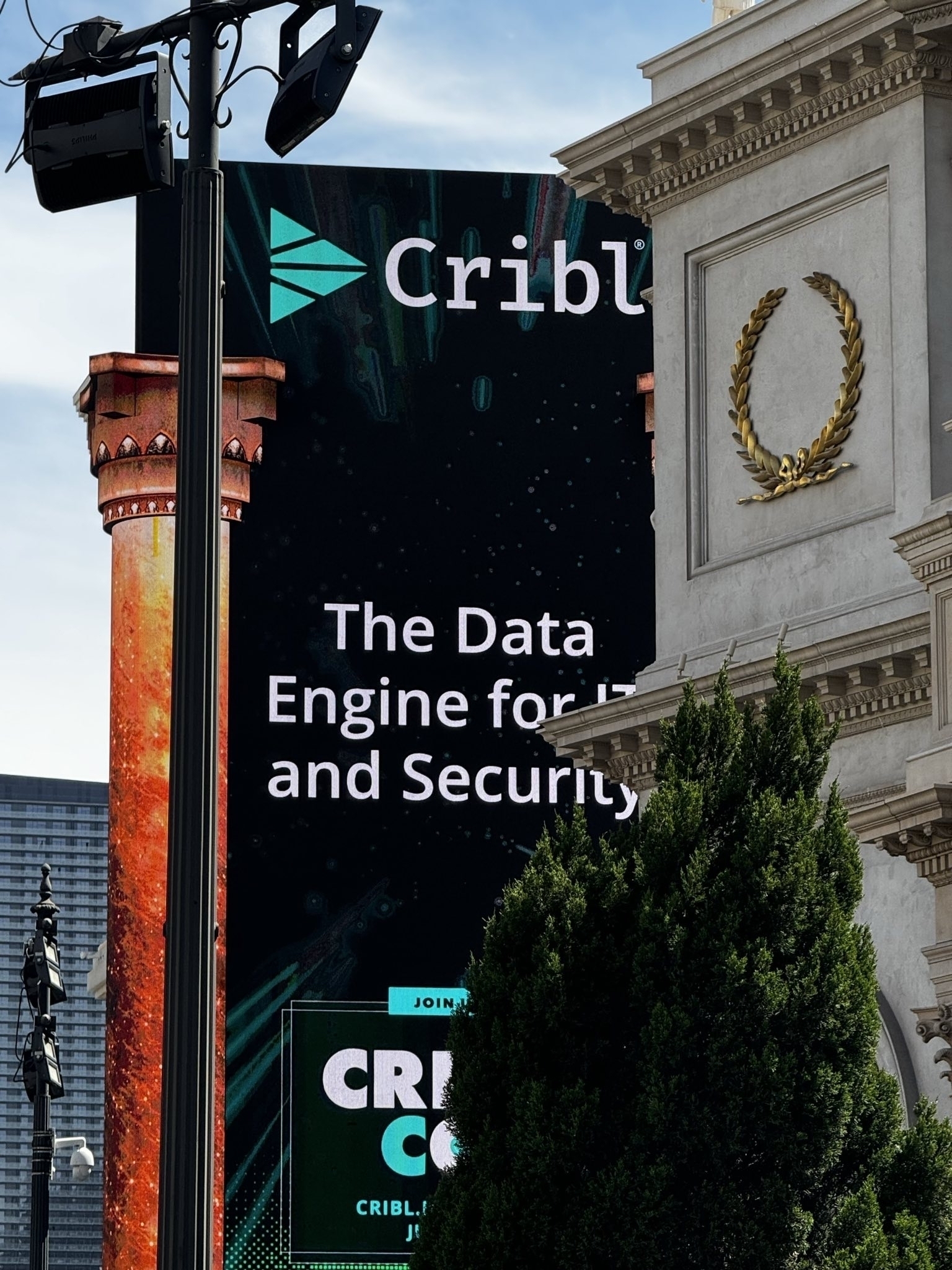 A large billboard advertising Cribl is prominently displayed beside a classical-style building adorned with a laurel wreath and foliage in the foreground.