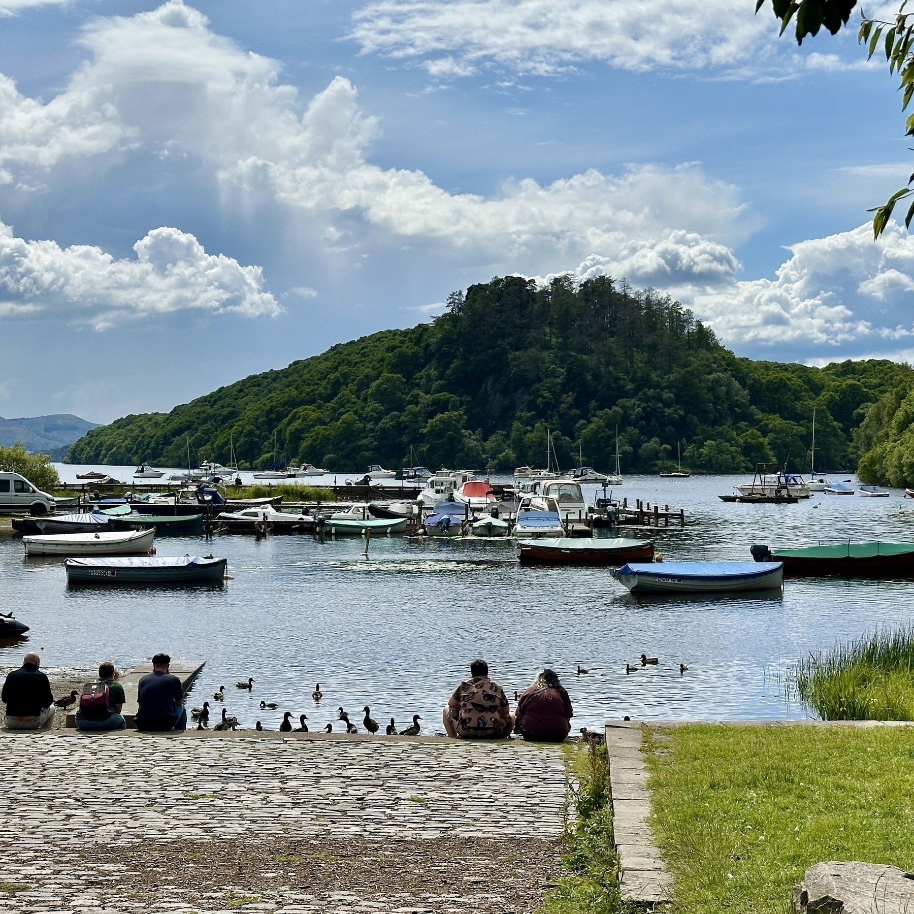 Auto-generated description: A picturesque scene of a lakeside area includes people sitting near the water, ducks swimming, boats anchored, and a lush green hill under a partly cloudy sky.