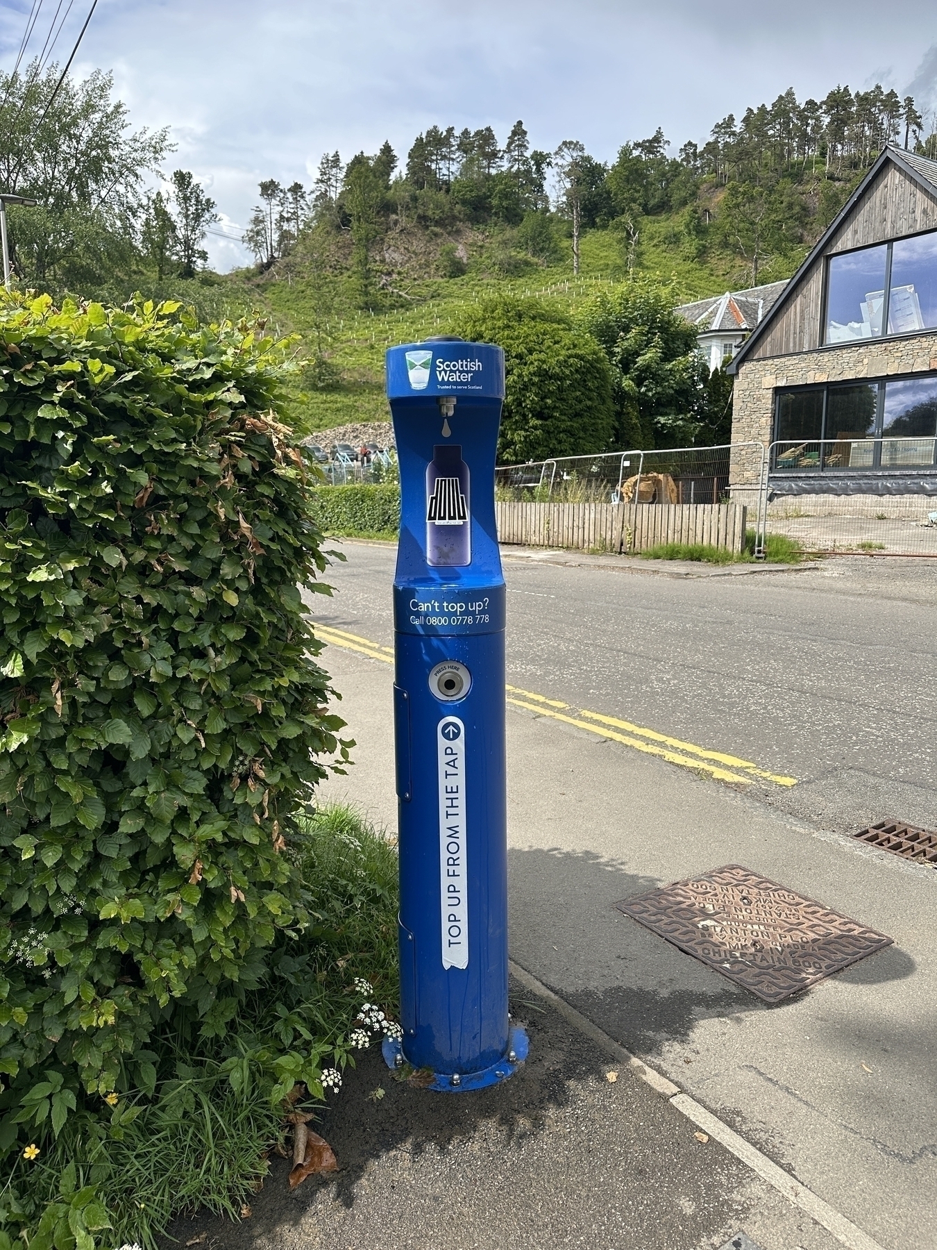 Auto-generated description: A blue water refill station is situated on a sidewalk beside a road, with a hillside and buildings in the background.