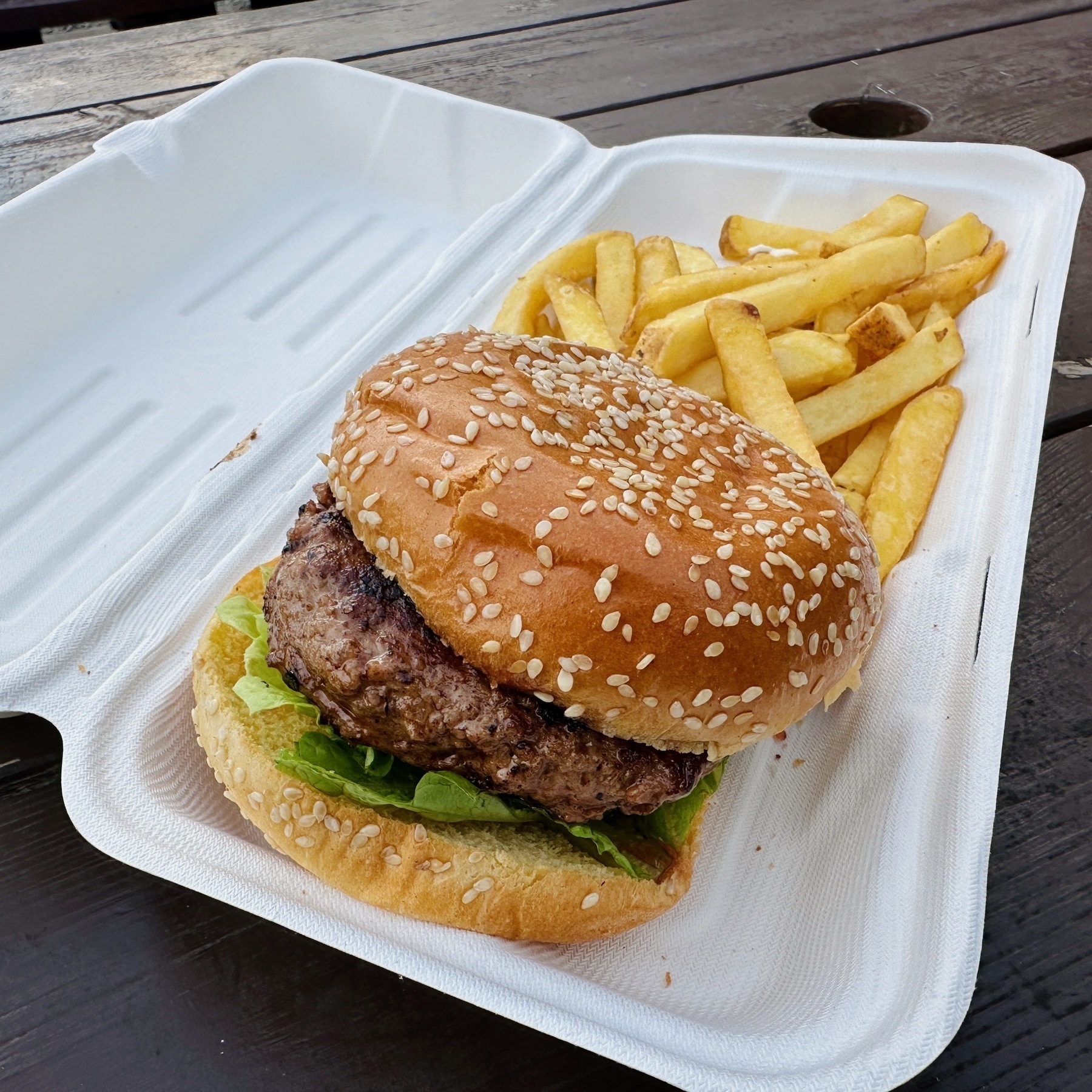 Auto-generated description: A juicy sesame seed bun burger with lettuce, accompanied by a side of chips, sits in a white, open clamshell takeout container on a wooden table.