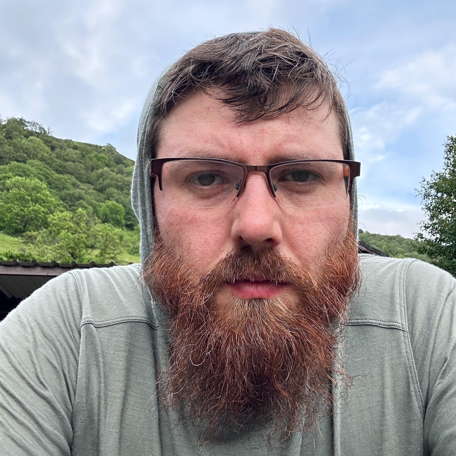 Auto-generated description: A bearded man with glasses is wearing a hooded jacket and standing outdoors with trees and hills in the background.