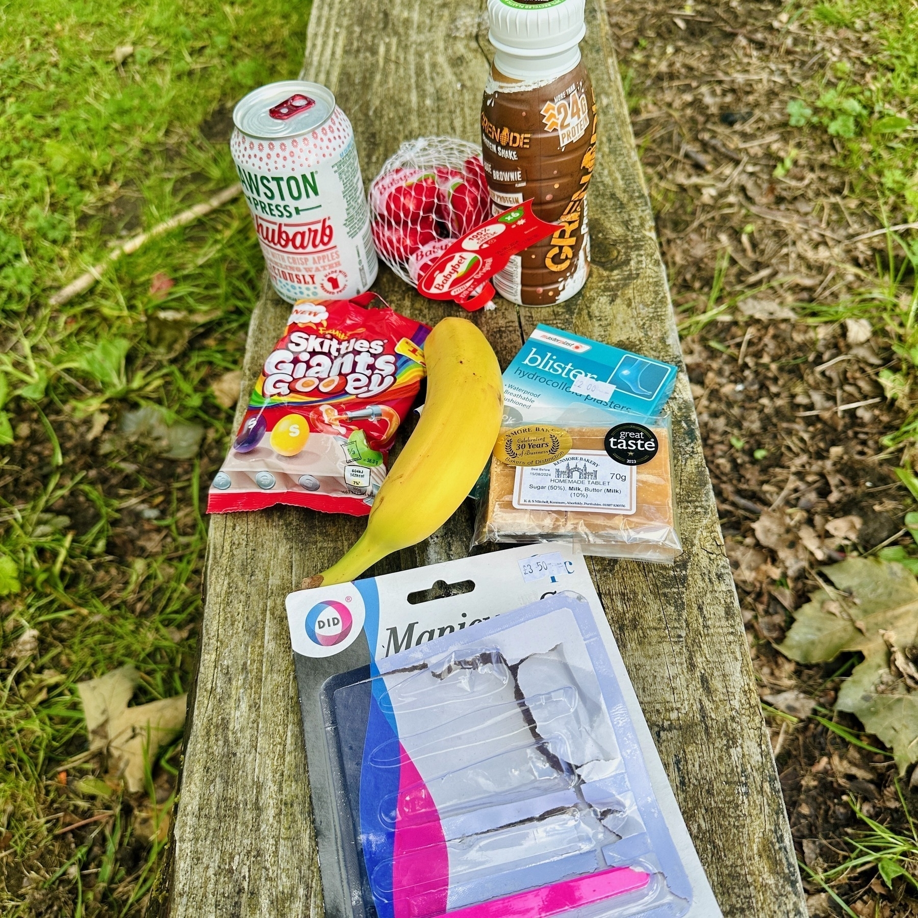 Auto-generated description: A selection of items including a can of rhubarb soda, a bottled drink, Skittles, a banana, a pack of blister plasters, a bag of snack cheese, and the packaging of a manicure set are displayed on a wooden surface outdoors.