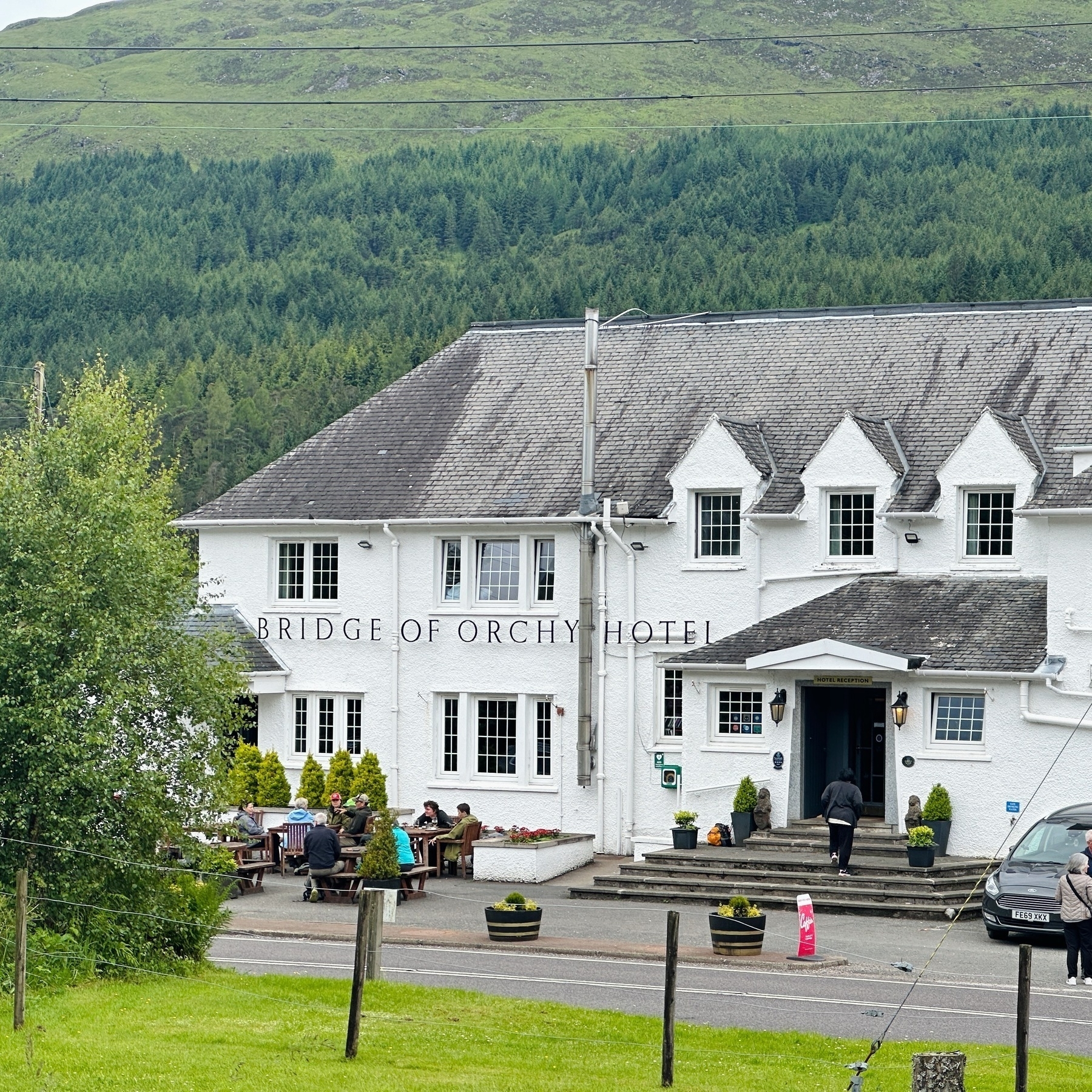 Auto-generated description: A white building labeled Bridge of Orchy Hotel sits against a backdrop of green hills, with people dining and a person walking towards the entrance.