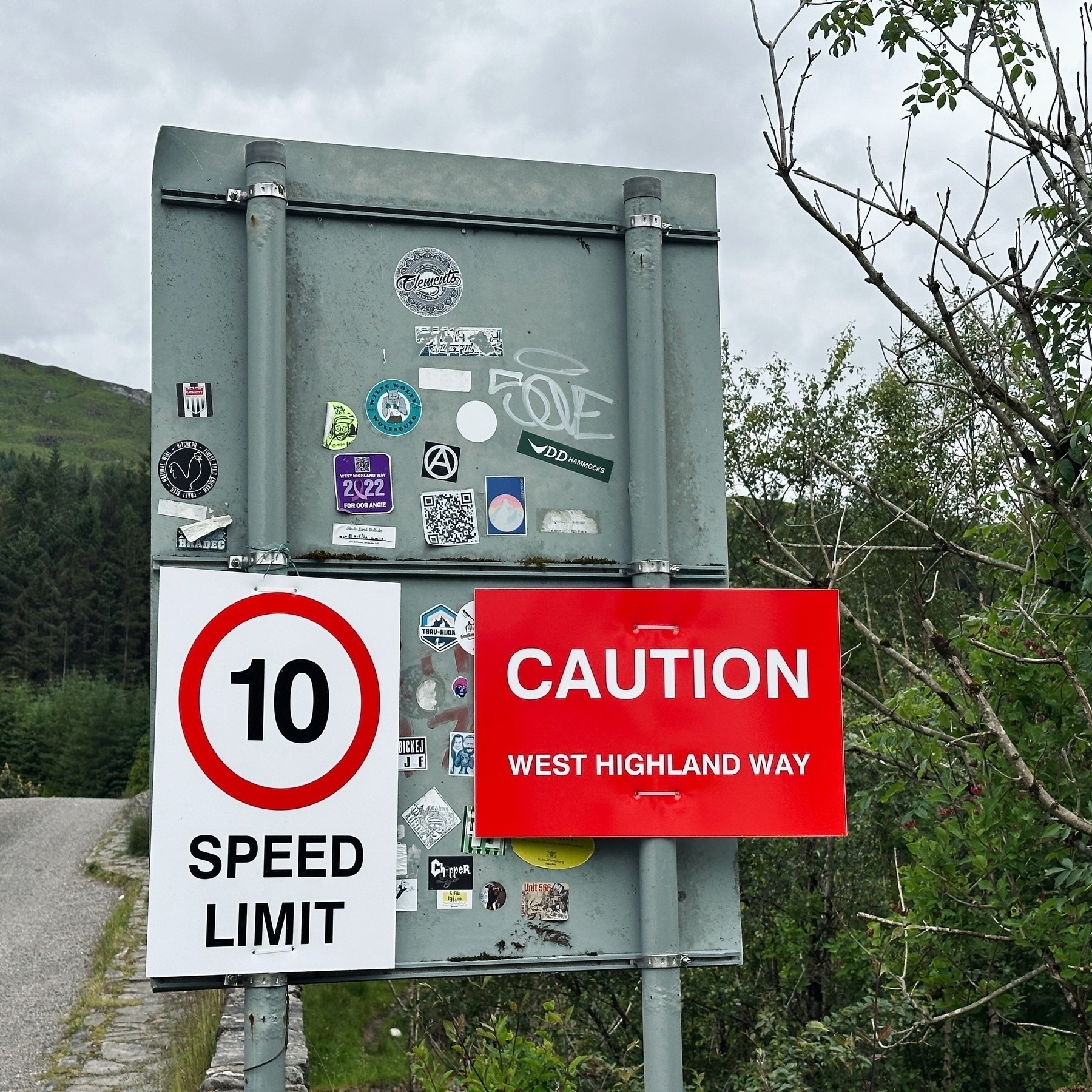 Auto-generated description: A sign on the side of a rural road indicates a 10 mph speed limit and cautions travelers about the West Highland Way, surrounded by various stickers and greenery in the background.