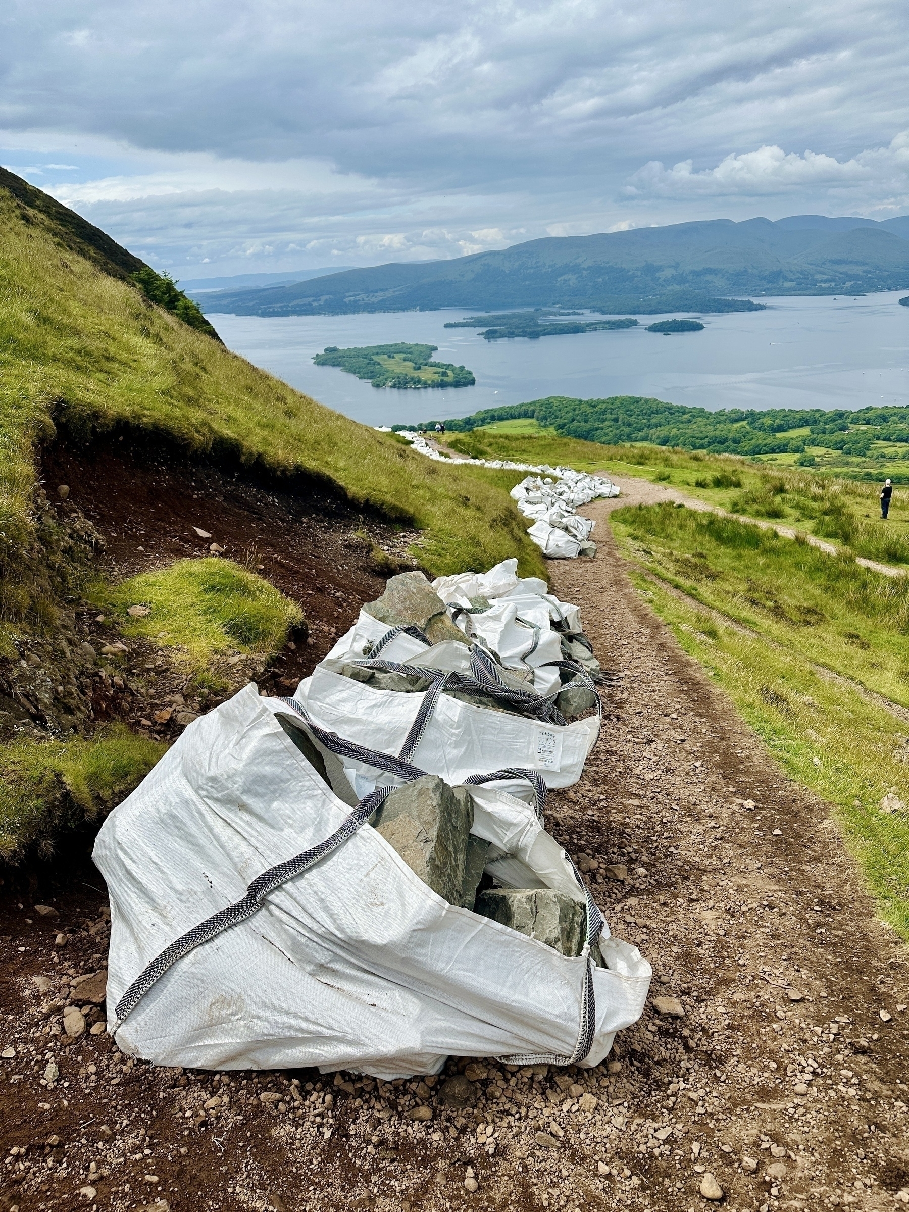 Large white bags filled with rocks are placed along a dirt path on a hill overlooking a scenic view of Loch Lomond and distant mountains.