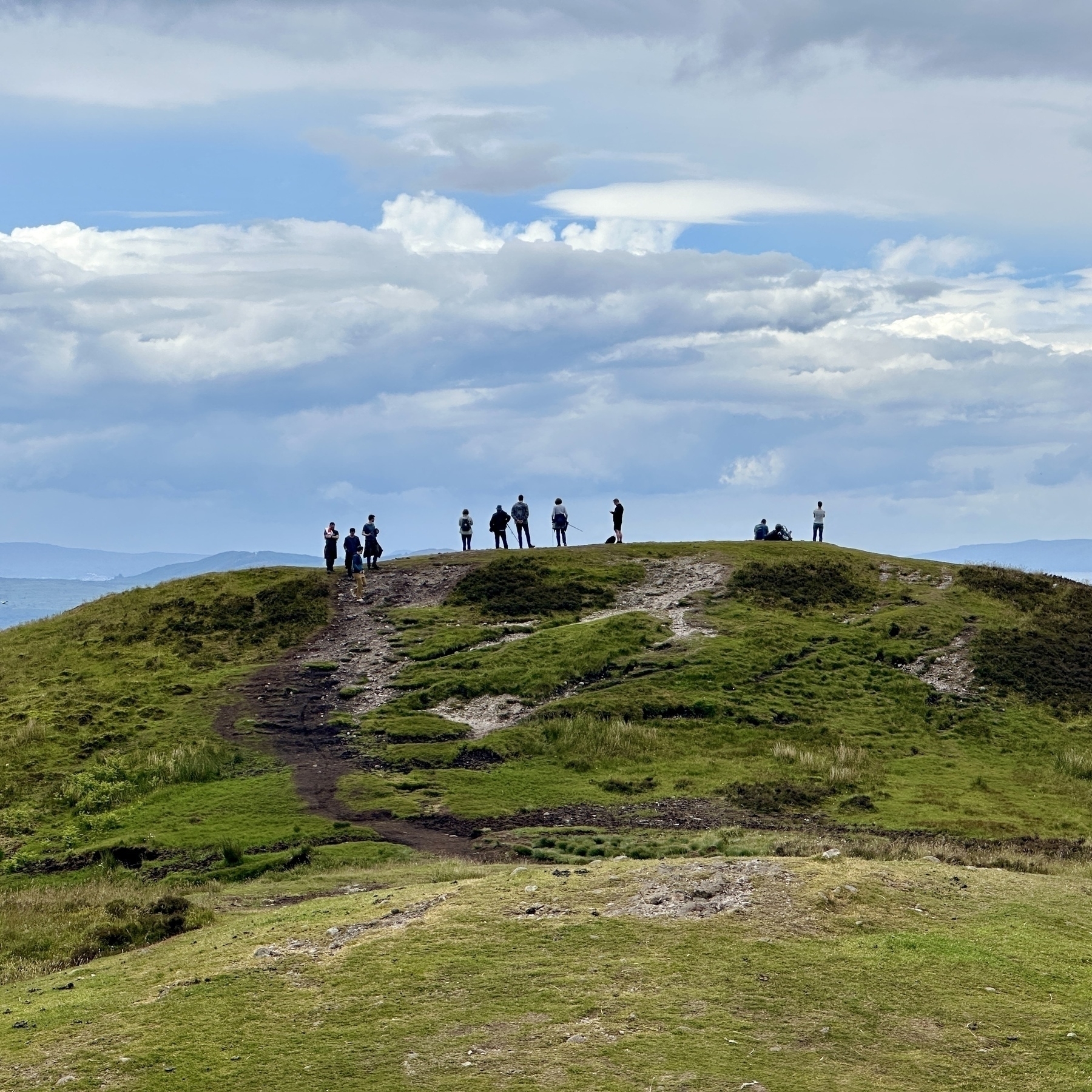 Auto-generated description: A group of people stand atop a grassy, gently sloping hill under a partly cloudy sky.