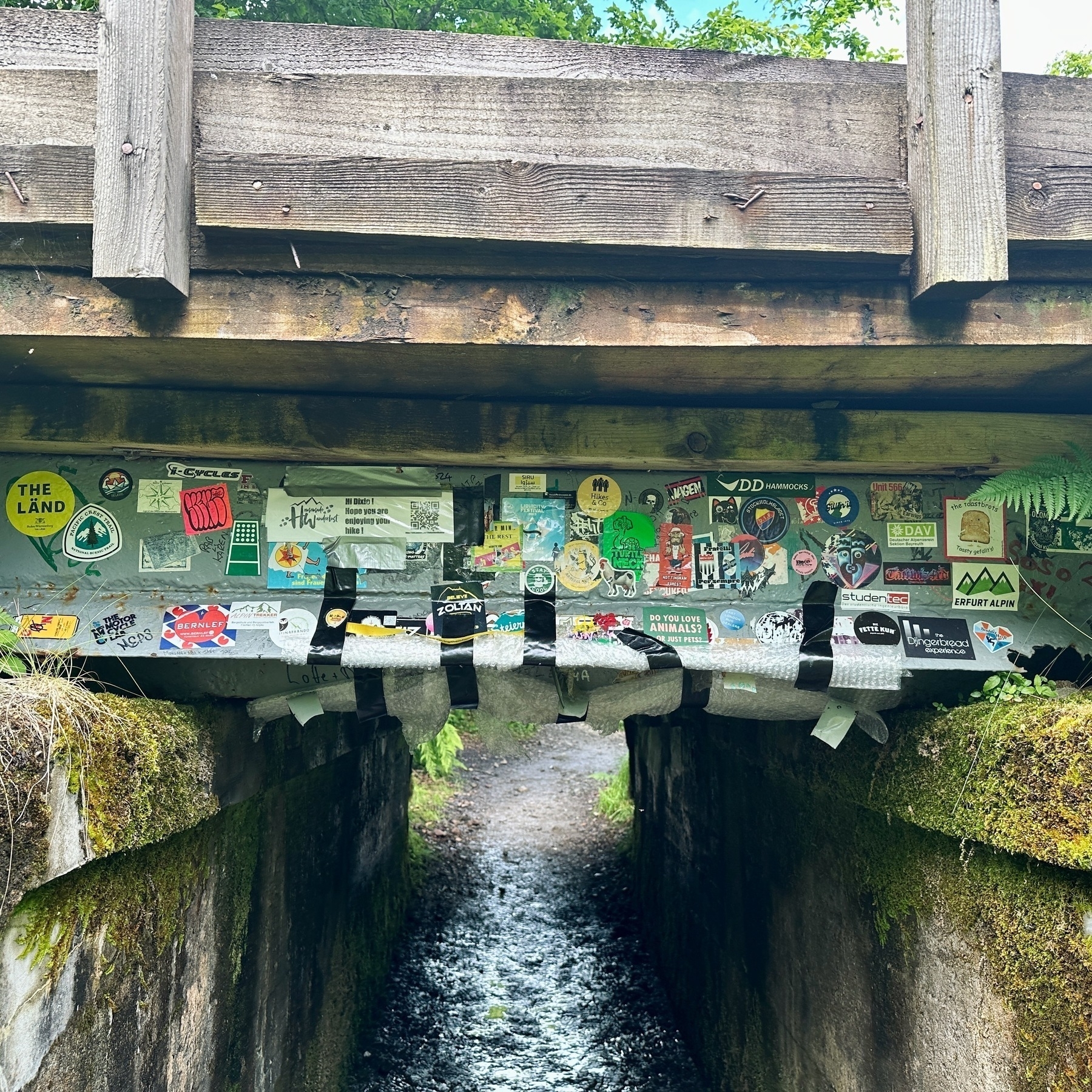 Auto-generated description: A small underpass beneath a wooden bridge is covered with a variety of colourful stickers and graffiti.