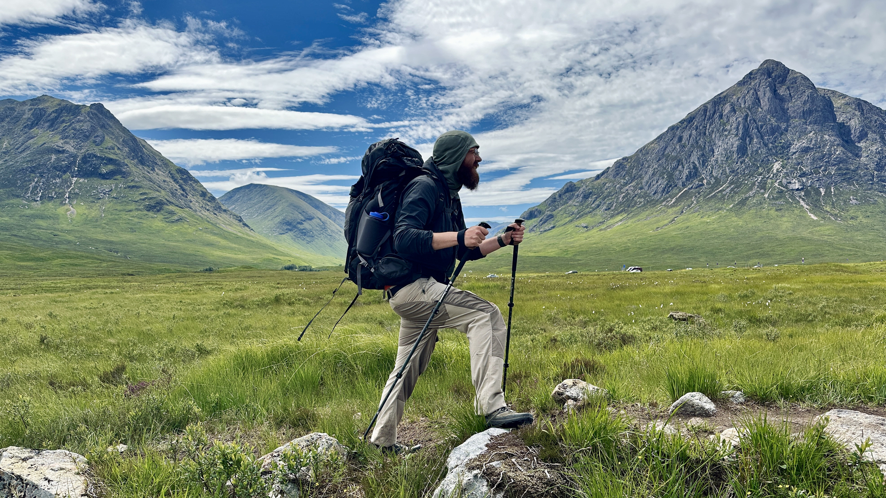 Auto-generated description: A person equipped with hiking gear walks through a vast, green mountainous landscape under a partly cloudy sky.