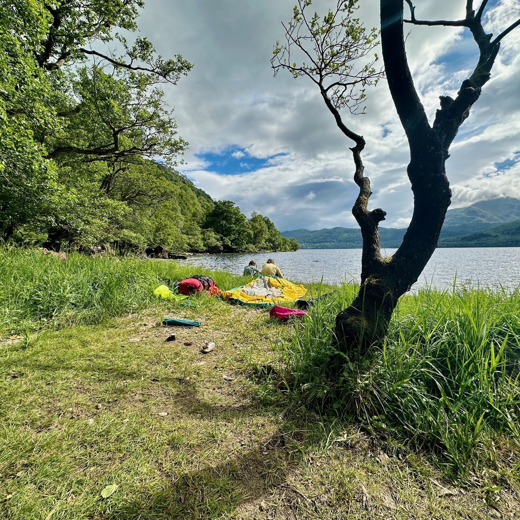 Auto-generated description: A lakeside scene with scattered camping gear near the shore, surrounded by trees and hills under a cloudy sky.