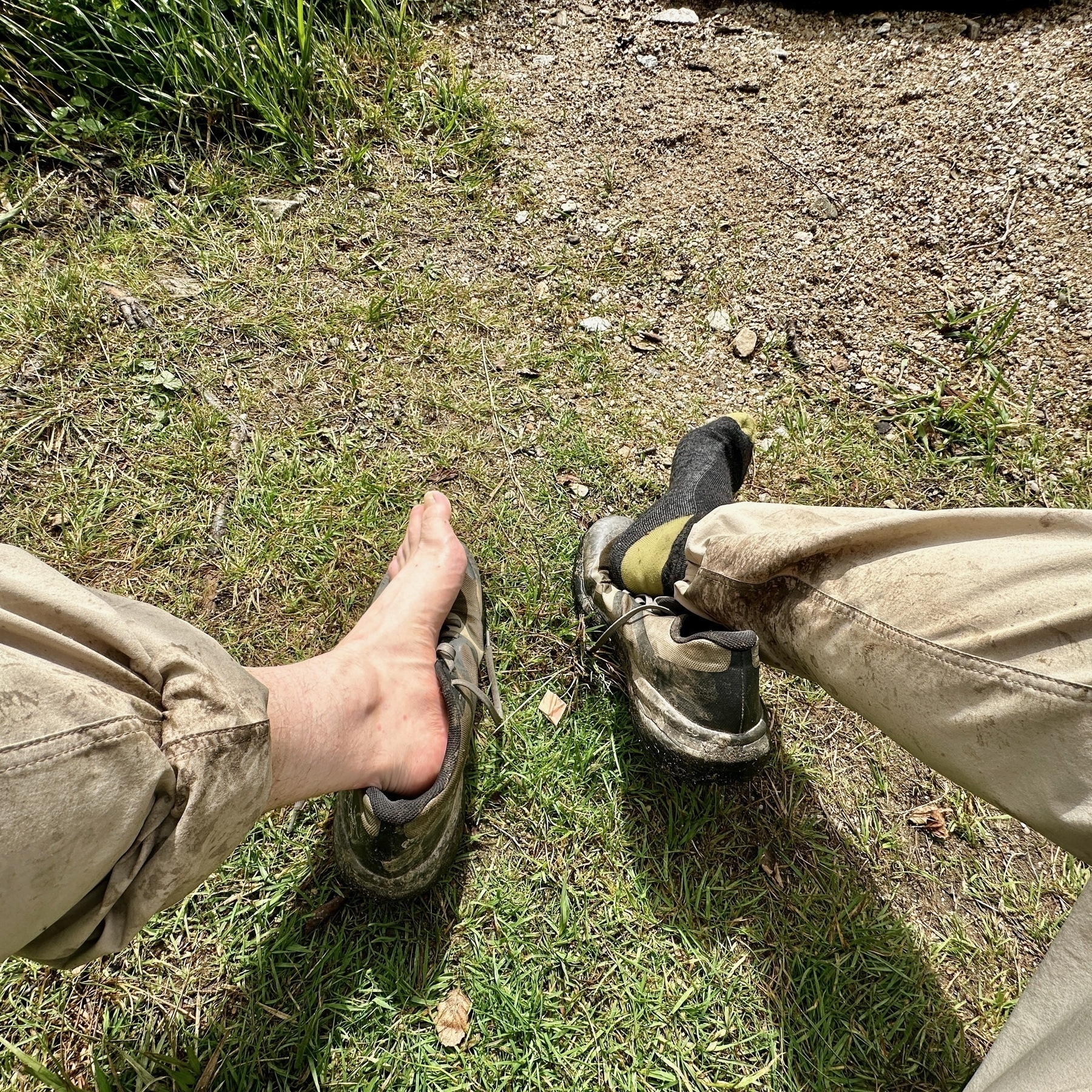 Auto-generated description: A person is sitting on grass with one foot out of a shoe, wearing dirty beige pants and resting near a rocky surface.