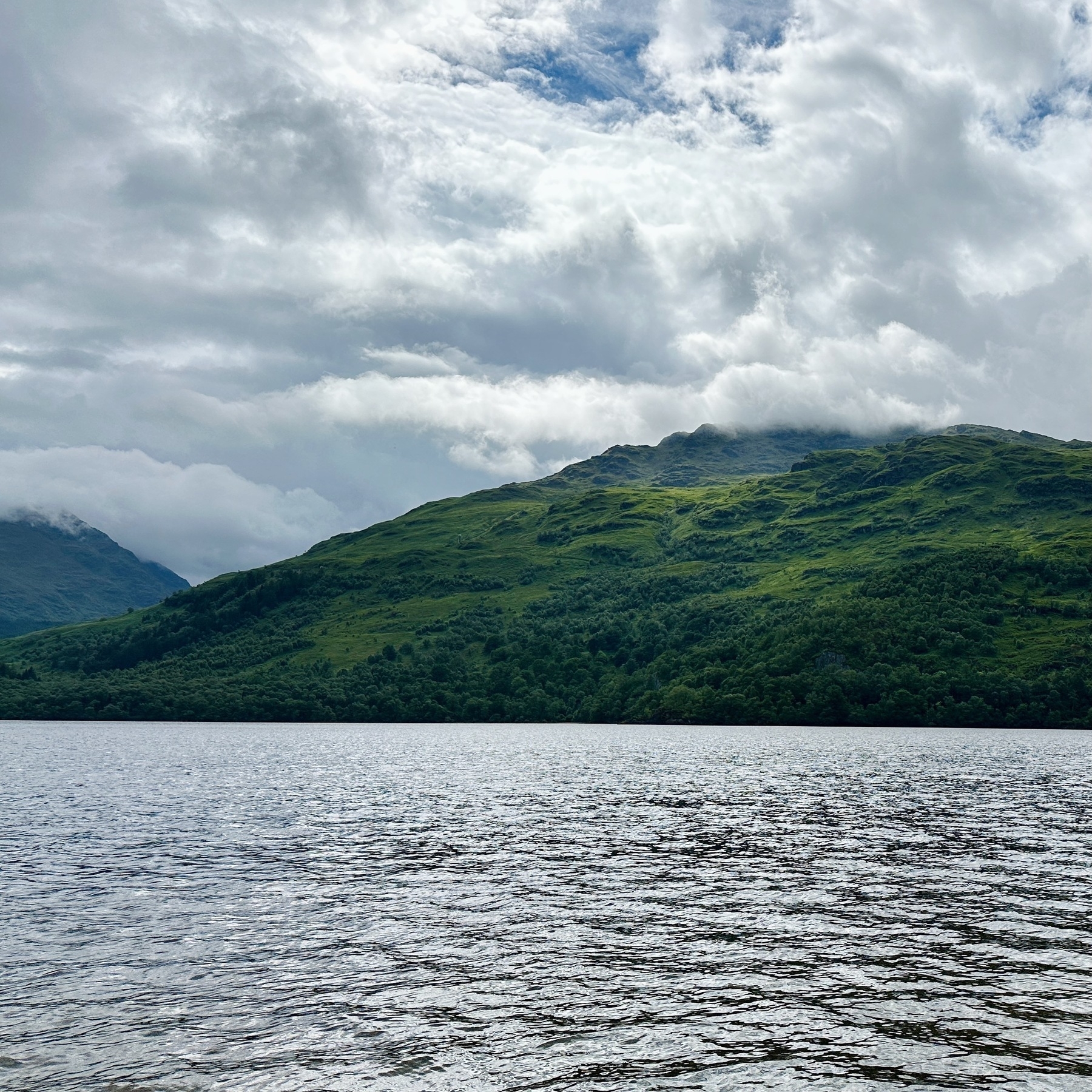 Auto-generated description: A serene landscape features a calm body of water in the foreground, with lush green hills and a cloudy sky in the background.