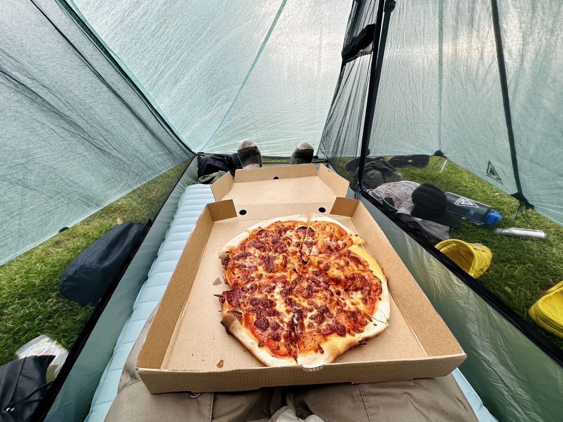 Auto-generated description: Inside a tent, a person enjoys a pizza placed on their lap while lying down, with camping gear visible alongside.