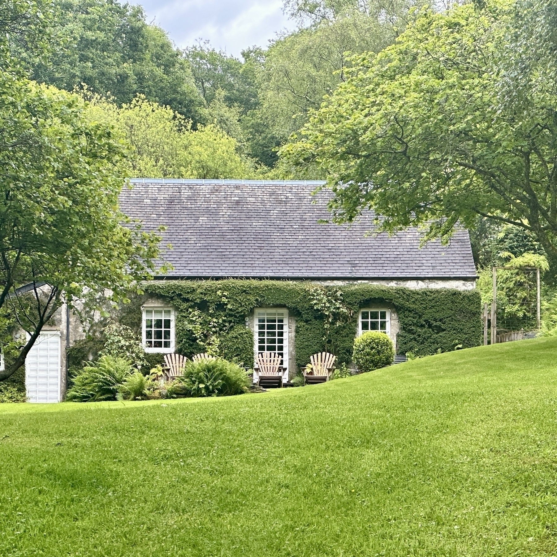 Auto-generated description: A charming stone cottage, covered in greenery, is nestled in a lush, well-manicured garden surrounded by tall trees.