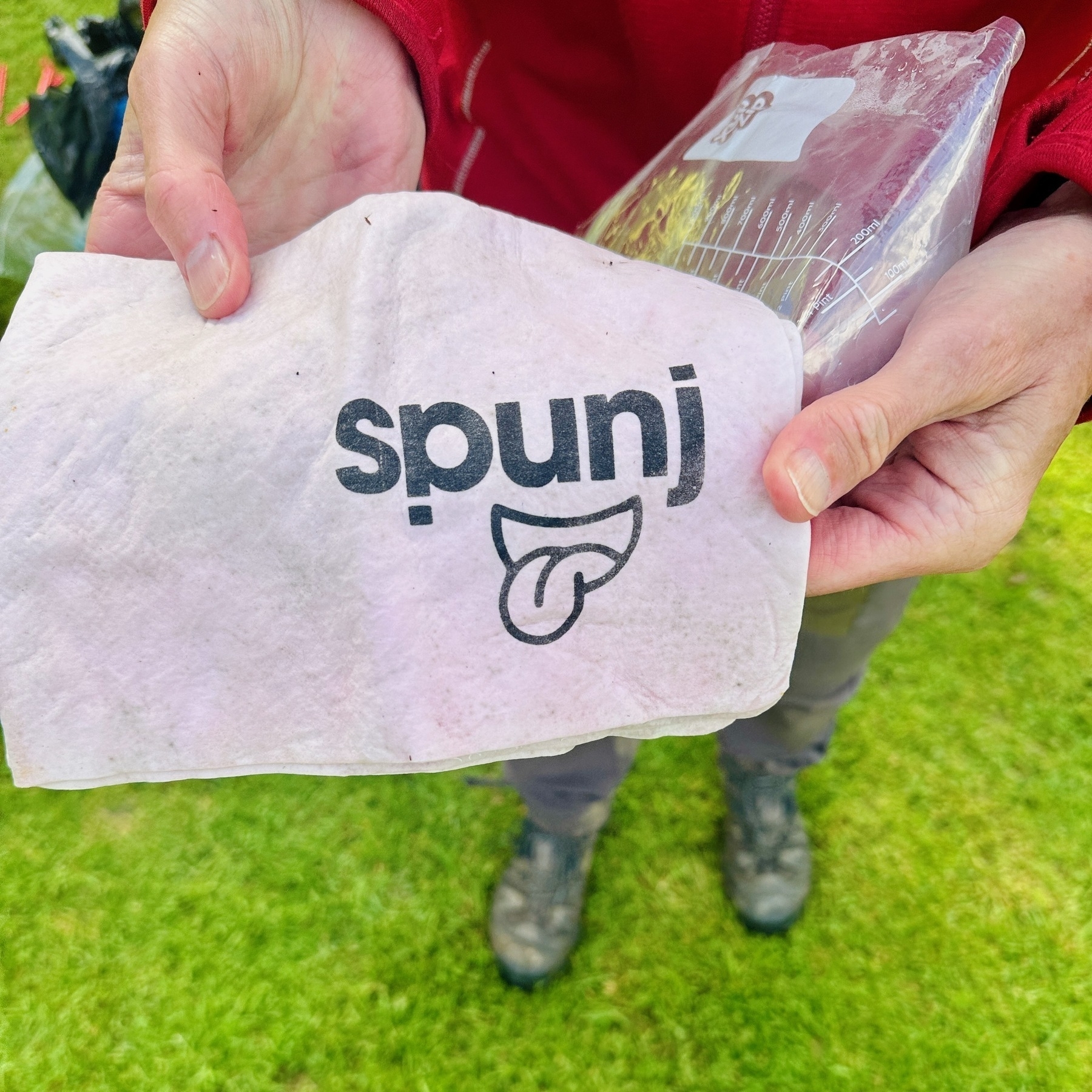 Auto-generated description: A person, seen from above, holds a sponge with the word Spunj and a smiling tongue logo, while standing on grass.