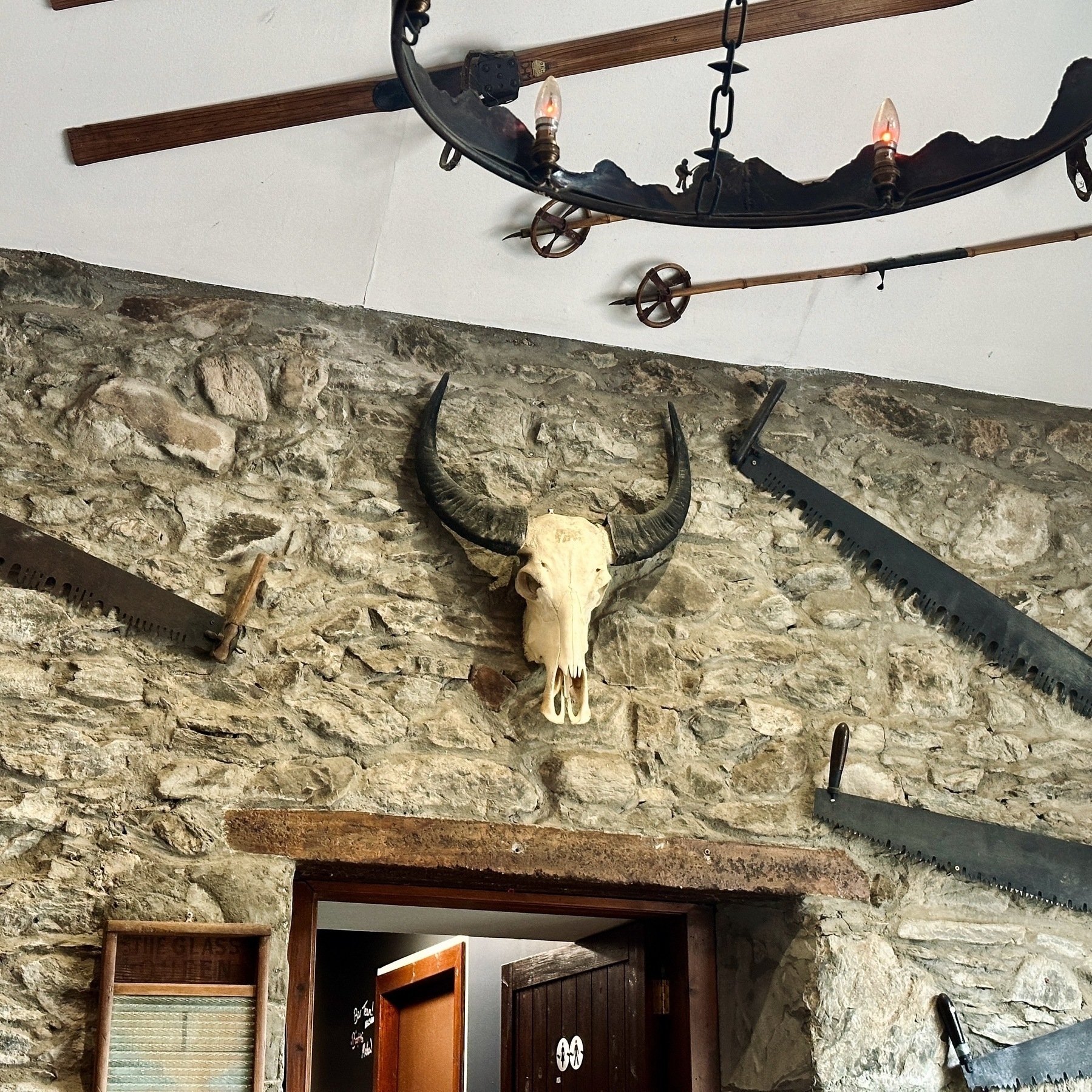 Auto-generated description: A rustic interior features a stone wall adorned with a mounted deer skull, old saw blades, and an iron chandelier.