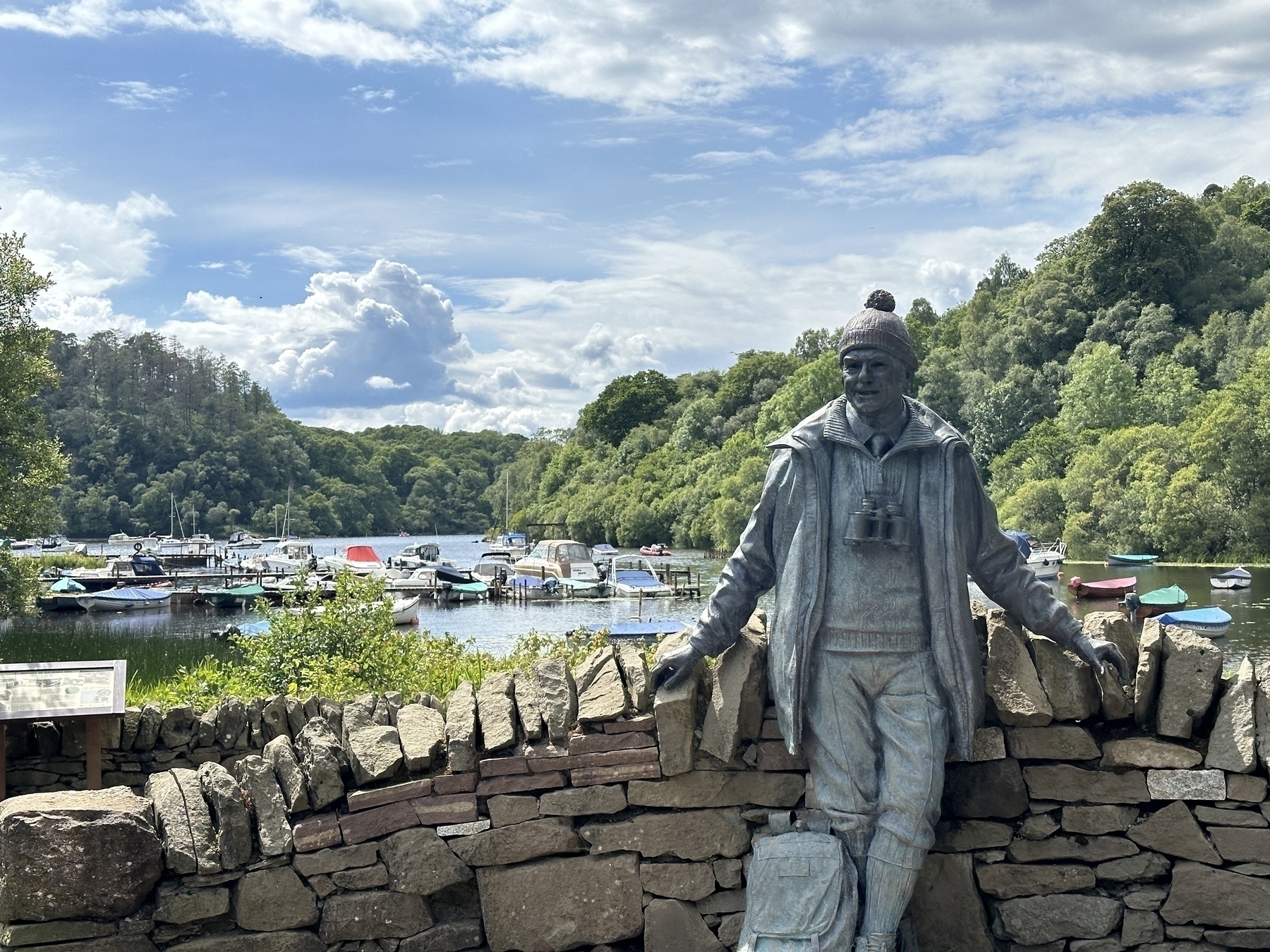 A statue of Tom Weir, dressed in outdoor attire, stands by a stone wall overlooking a scenic lake with boats and surrounded by lush greenery and wooded hills.